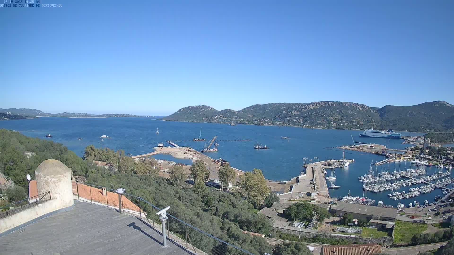 An elevated view of a harbor filled with boats, under a clear blue sky, shows ongoing construction near a ferry and a large body of water surrounded by hills.