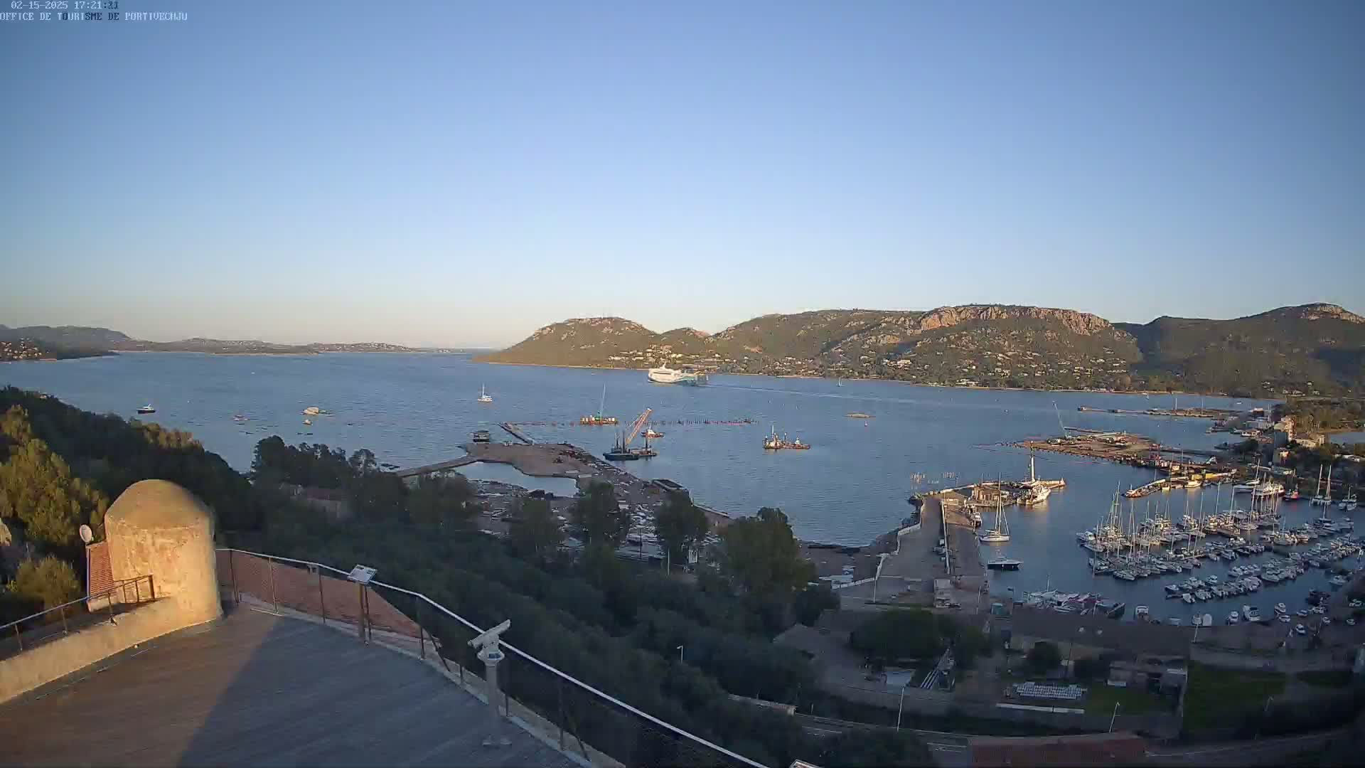 An elevated view of a calm bay with numerous boats in a marina, a large ship in the distance, and hills under a clear blue sky.