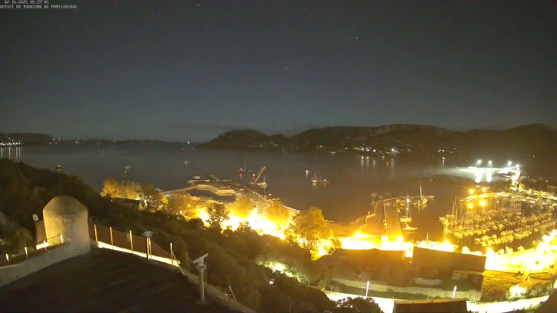 A nighttime view of a harbor filled with boats, illuminated by lights, nestled between hills under a clear, starry sky.