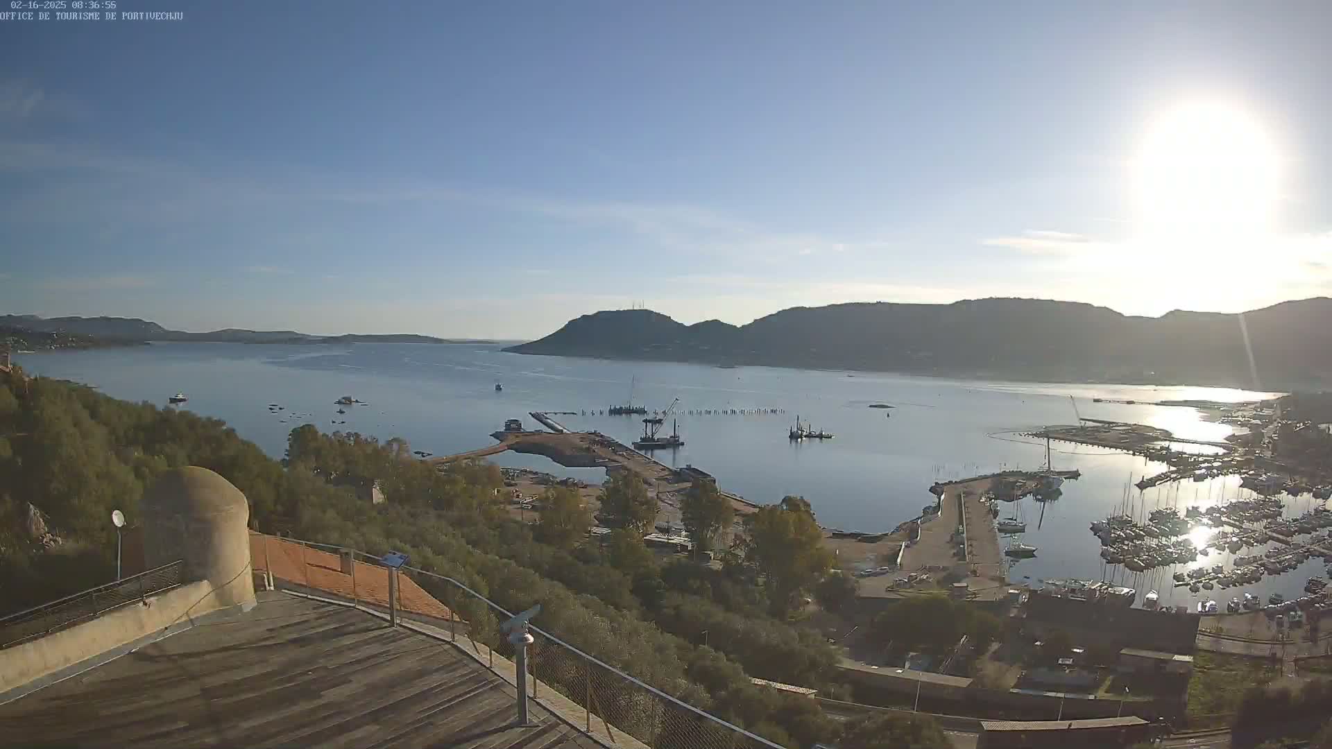 A sunny, high-angle view of a harbor with numerous boats and construction activity, nestled between hills and a calm body of water.
