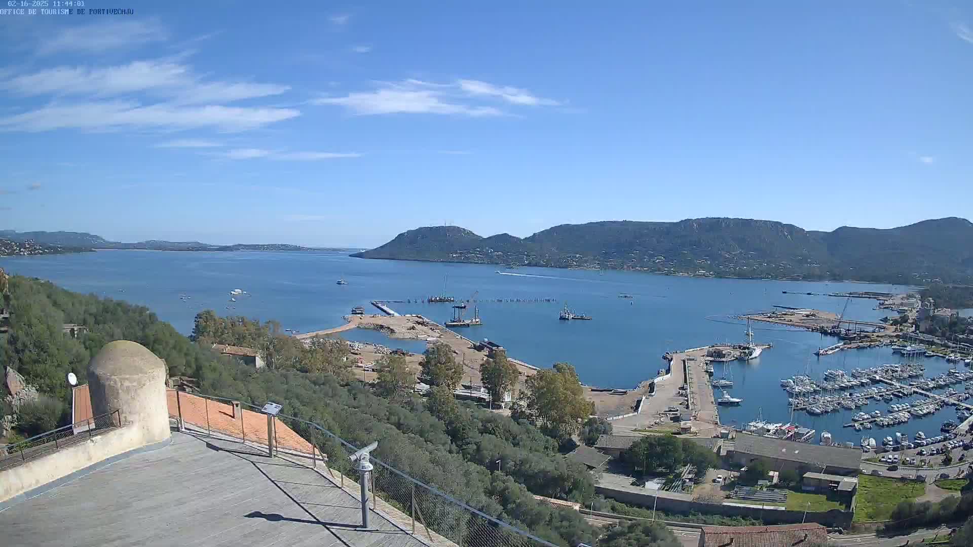 A high-angle view of a harbor filled with boats, under a clear blue sky with a few wispy clouds, situated between hills and a large body of water.