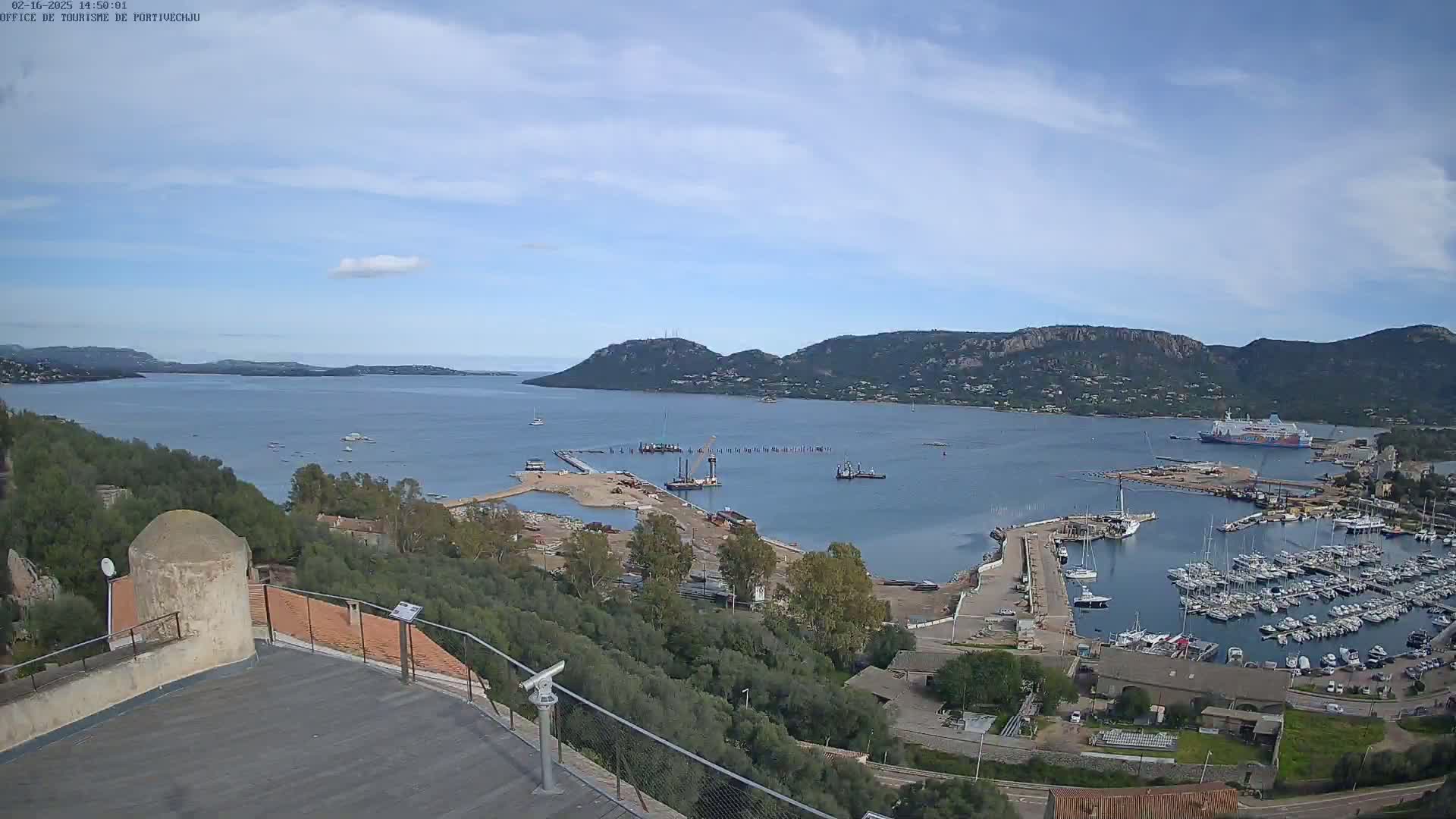 A panoramic view from a high vantage point shows a harbor filled with boats, construction activity near a pier, and a large body of water surrounded by hills under a mostly clear sky.