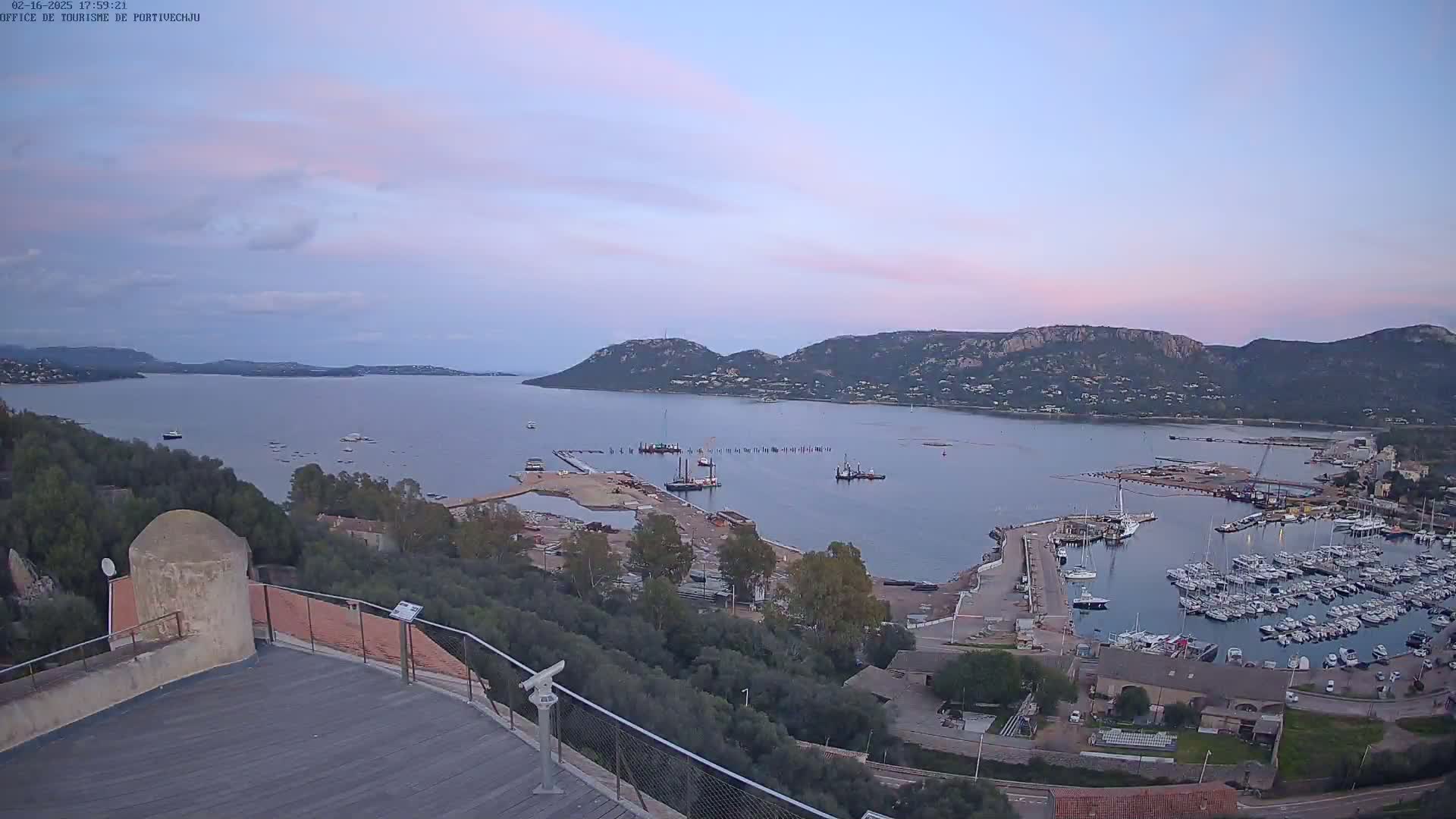 A high-angle, twilight view of a harbor filled with boats, construction activity, and a calm bay surrounded by hills under a pale pink and lavender sky.