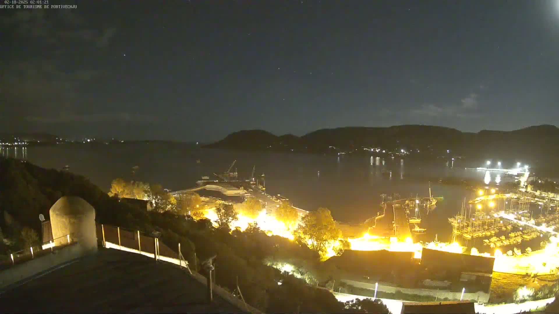 A nighttime view of a harbor filled with boats, illuminated by lights, under a mostly clear sky with a few faint clouds.