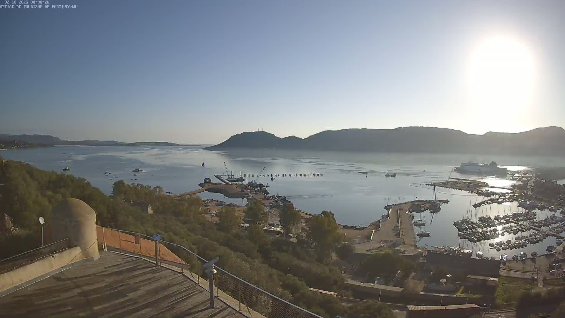 A high-angle, sunny view of a harbor filled with boats, a shipyard with construction equipment, and a calm body of water surrounded by hills and greenery under a clear sky.