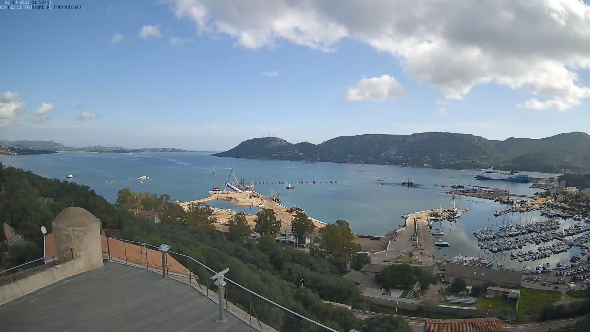 A partly cloudy day overlooks a harbor with many boats and a construction site, nestled between hills and a calm bay.