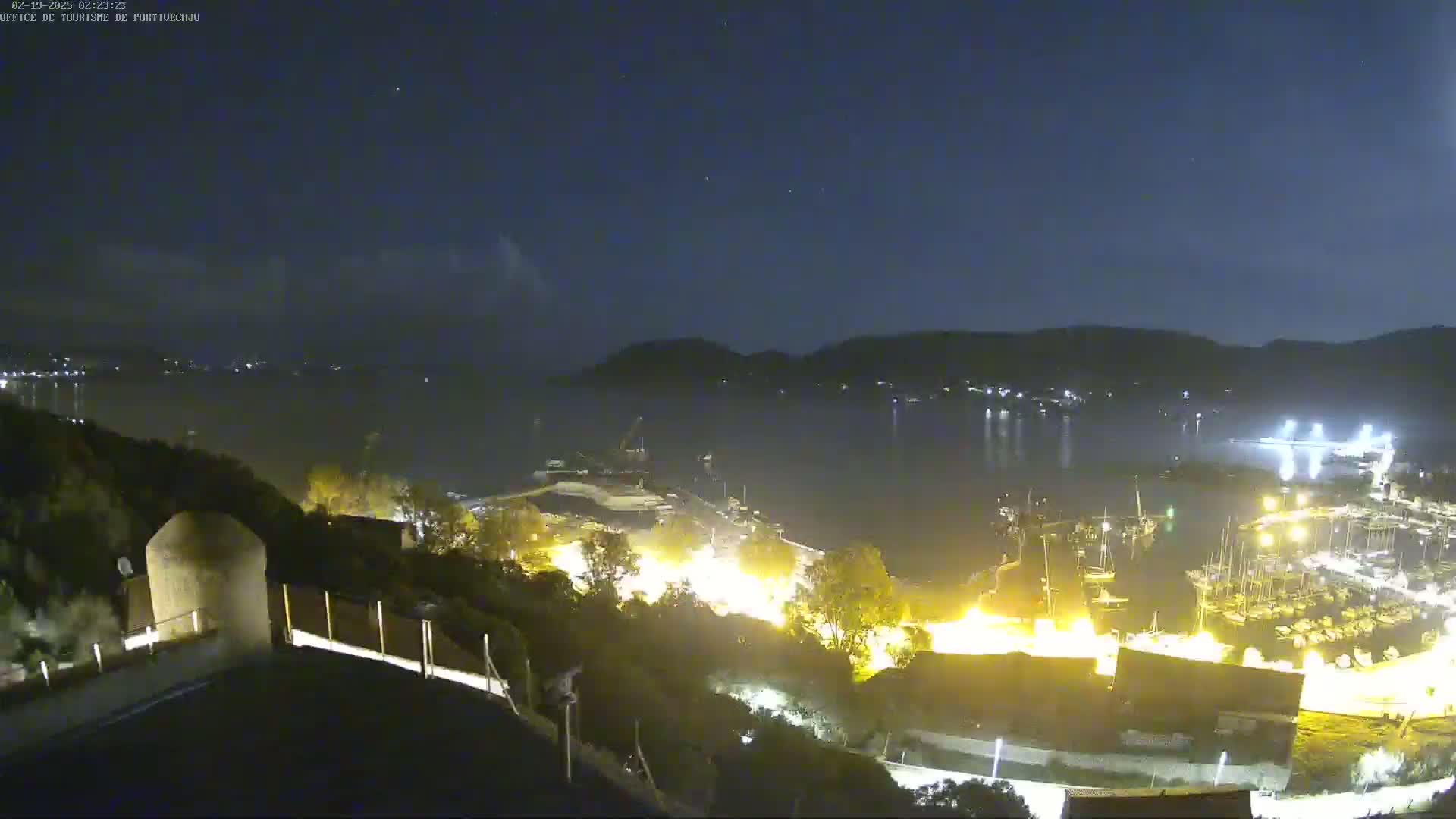 A nighttime view of a harbor filled with boats, illuminated by lights, and a dark body of water under a mostly clear sky.