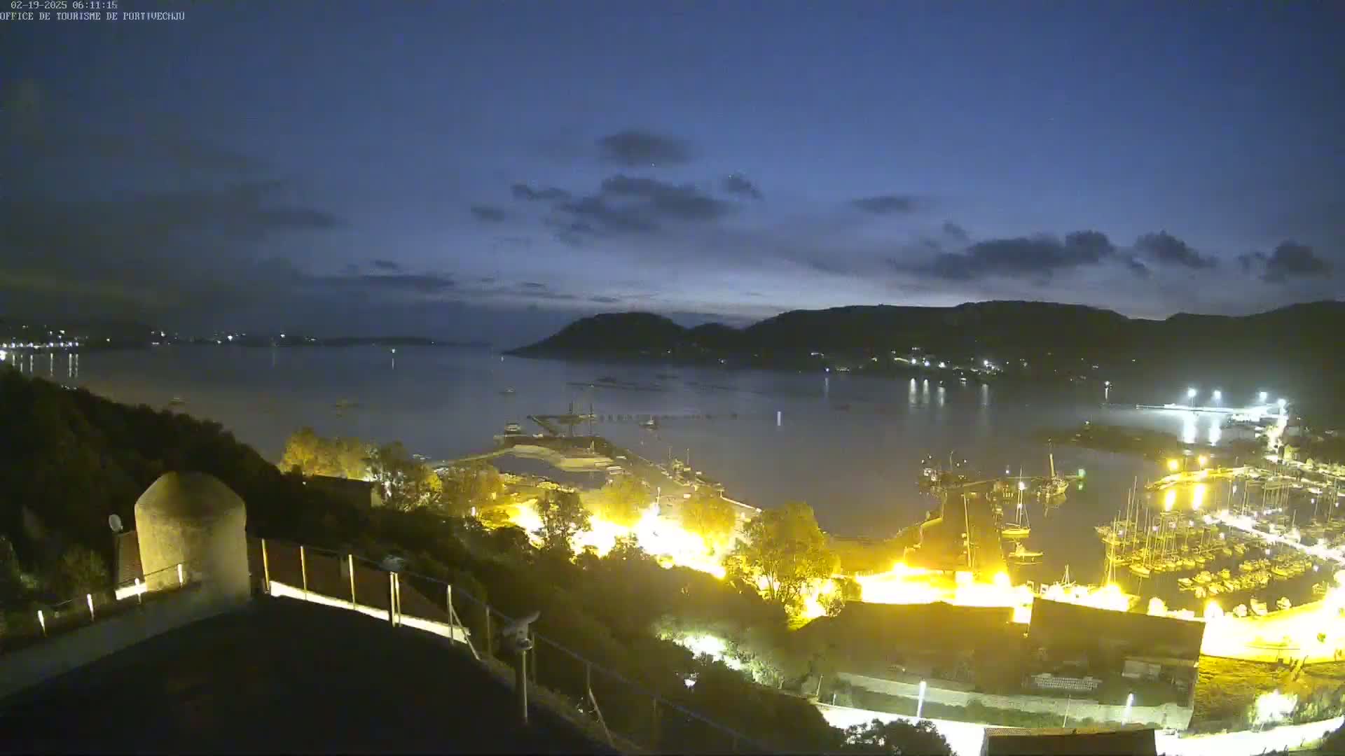 A nighttime, coastal scene shows a calm bay with a marina filled with boats, illuminated by artificial lights under a partly cloudy, dark blue sky.