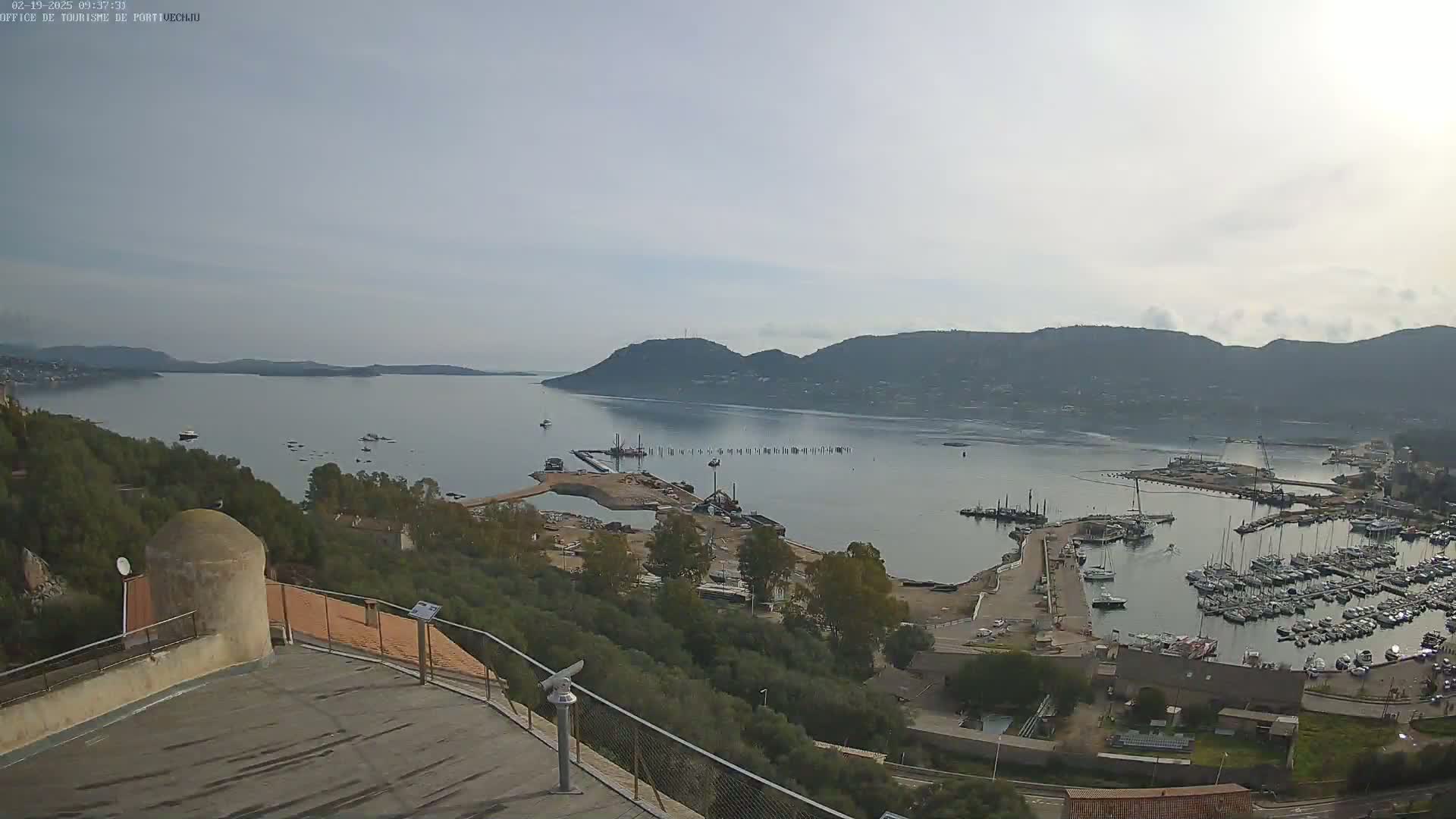 A high-angle, long shot shows a calm bay with many boats docked in a marina, surrounded by hills and green vegetation under a mostly clear sky.
