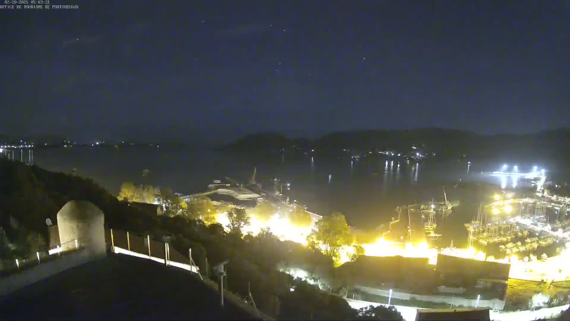 A nighttime view shows a harbor filled with boats, illuminated buildings, and dark hills under a clear, starry sky.