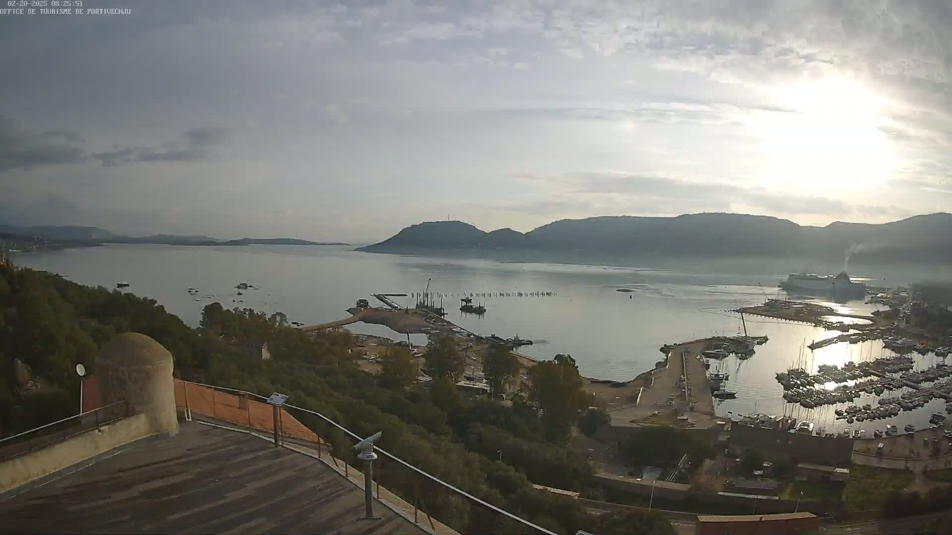 An elevated view shows a calm bay with a marina filled with boats, a large ferry, and distant mountains under a mostly cloudy sky.