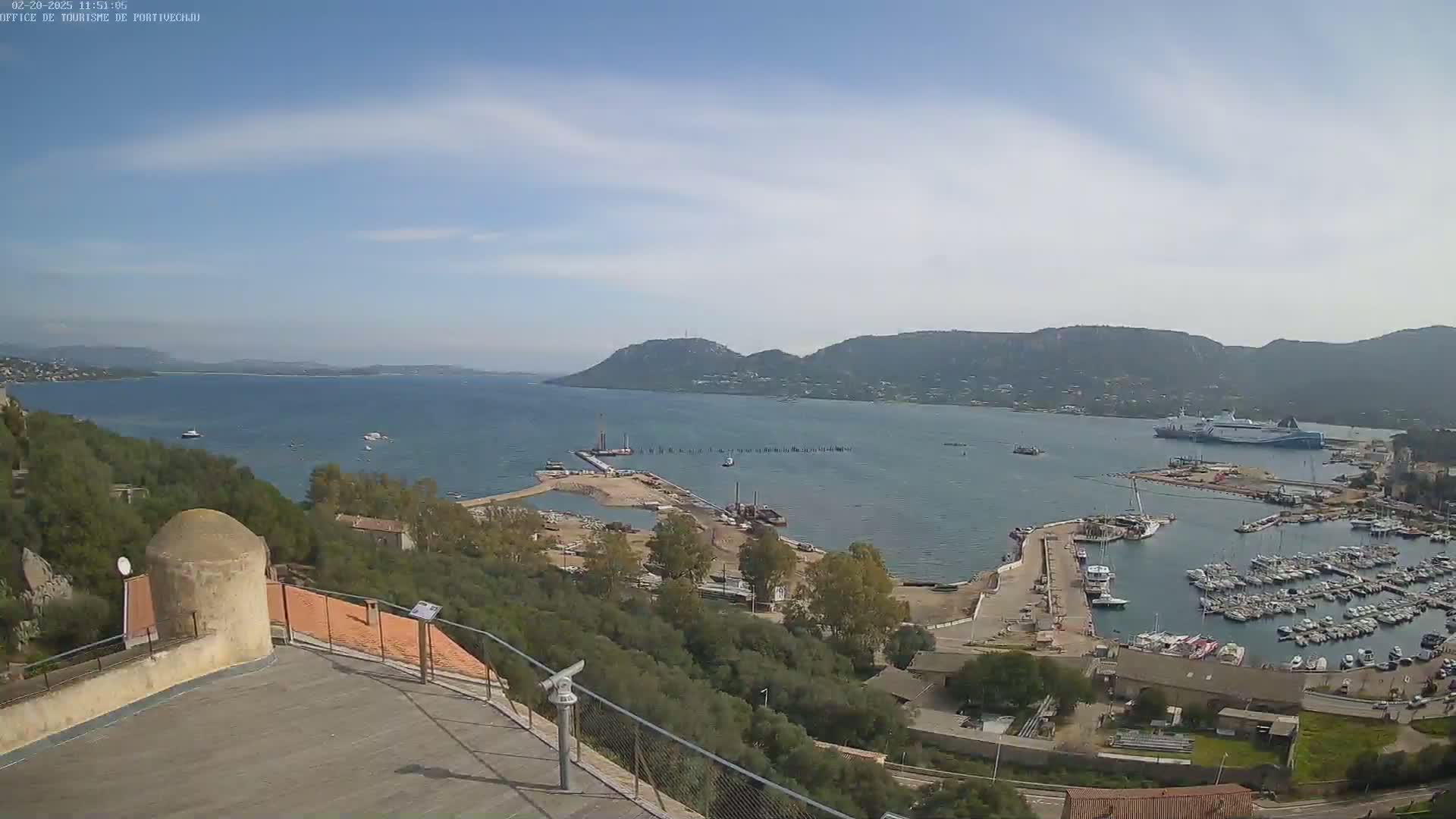 A high-angle, long shot shows a harbor filled with boats, docked alongside a shoreline with low-lying hills and a calm body of water under a mostly clear sky.