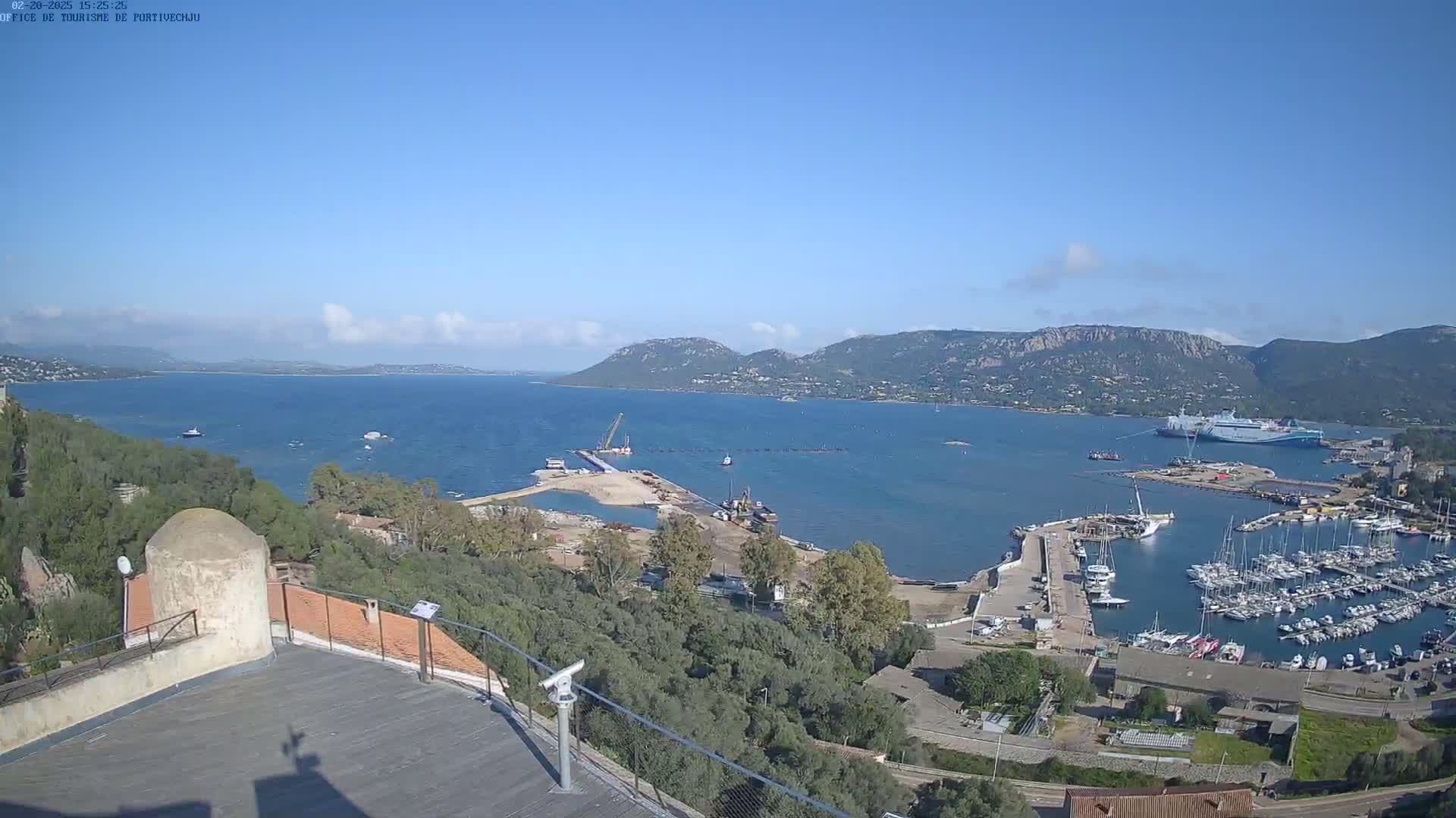 A mostly clear day reveals a harbor filled with boats, construction underway near a shoreline, and mountains in the distance, all viewed from a high vantage point.