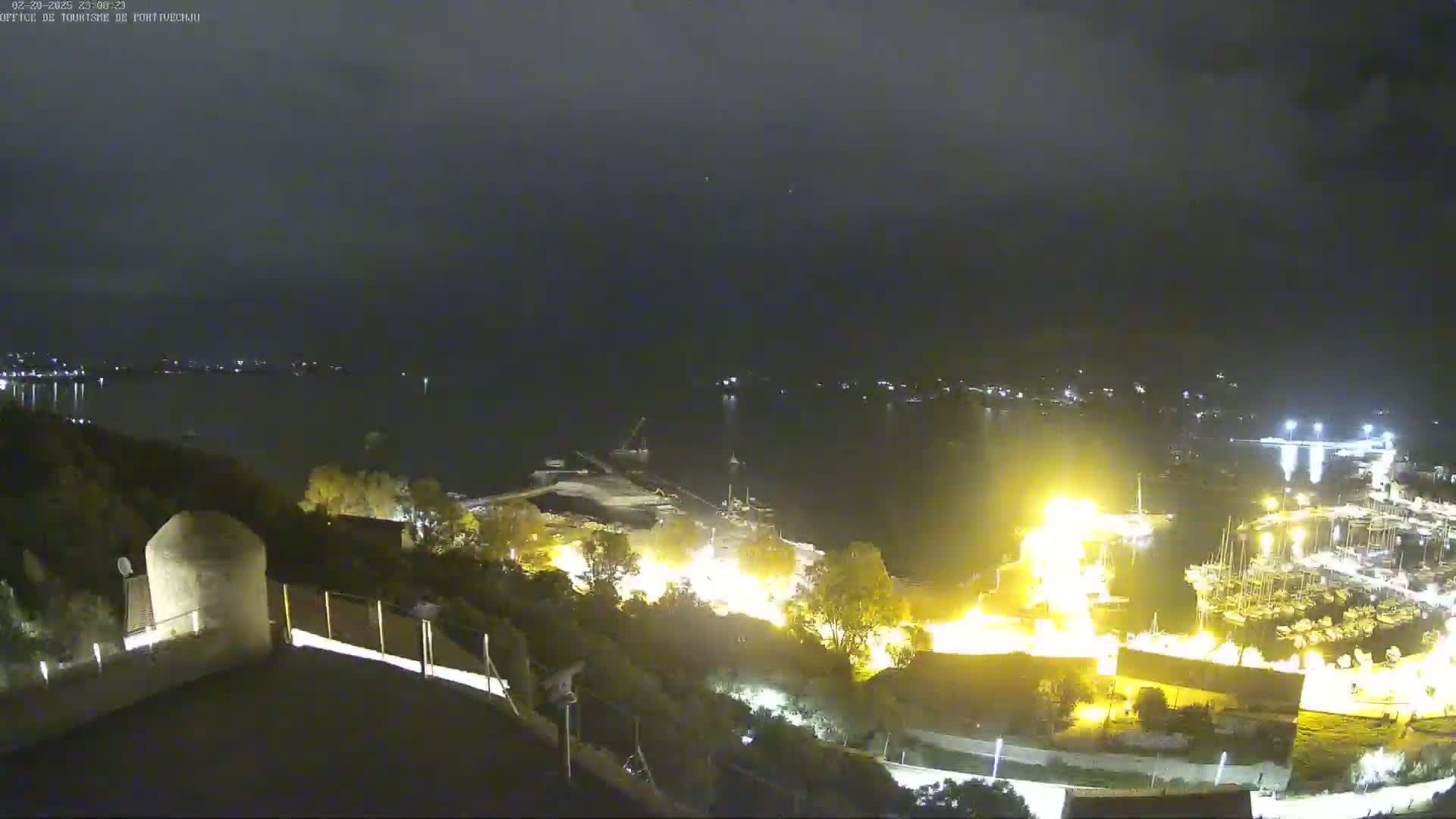 A nighttime, high-angle view of a harbor filled with boats, illuminated by lights, with a dark, calm body of water extending into the distance under a mostly clear sky.
