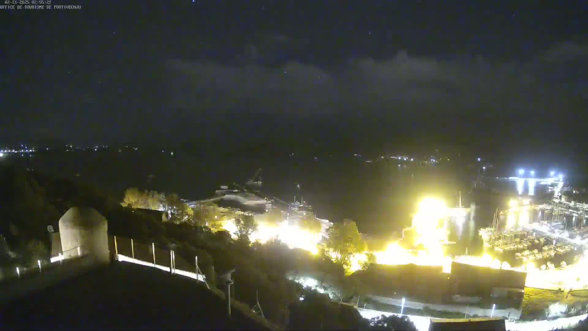 A nighttime, high-angle view shows a waterfront town with bright lights and a large fire or intense light source near a marina, under a partly cloudy sky.