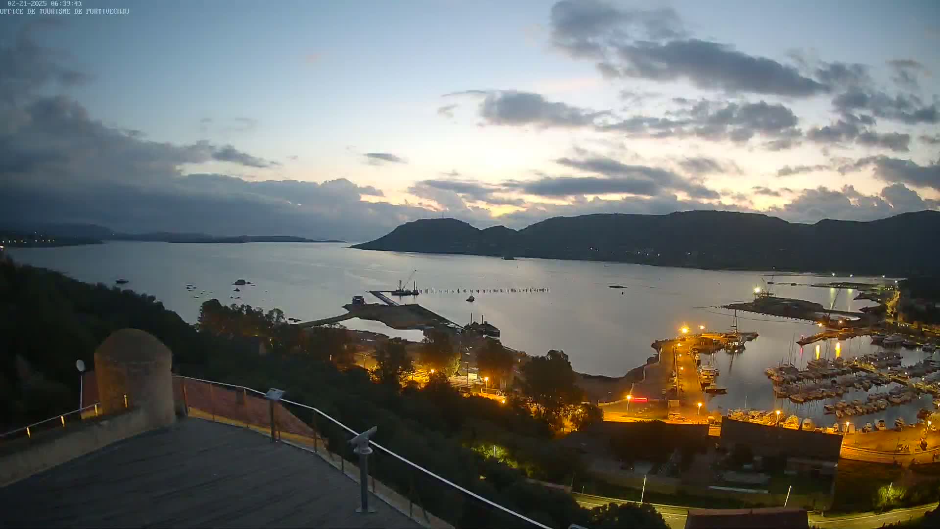 An elevated nighttime view of a calm bay with a marina filled with boats, under a partly cloudy sky, shows hills in the background.