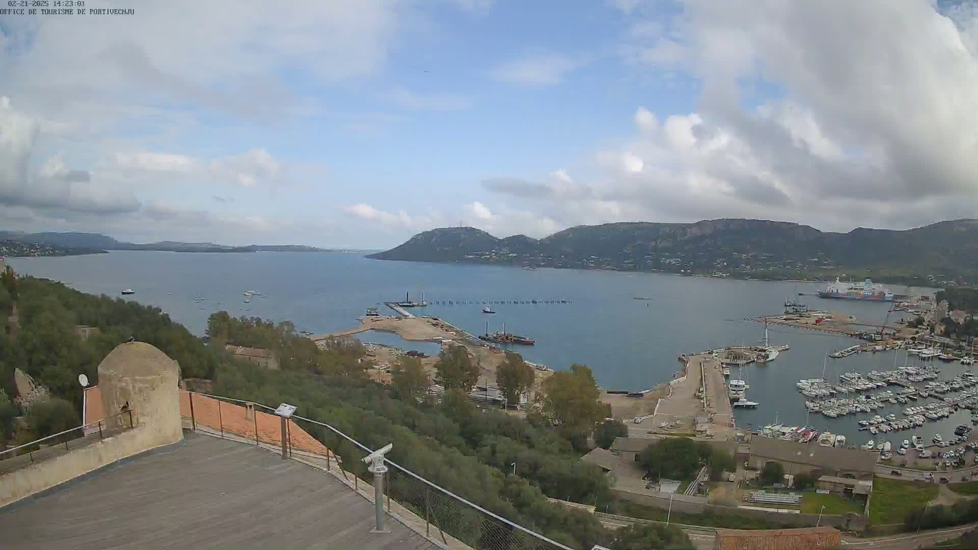 A high-angle view of a harbor filled with boats and yachts, nestled between hills under a partly cloudy sky.