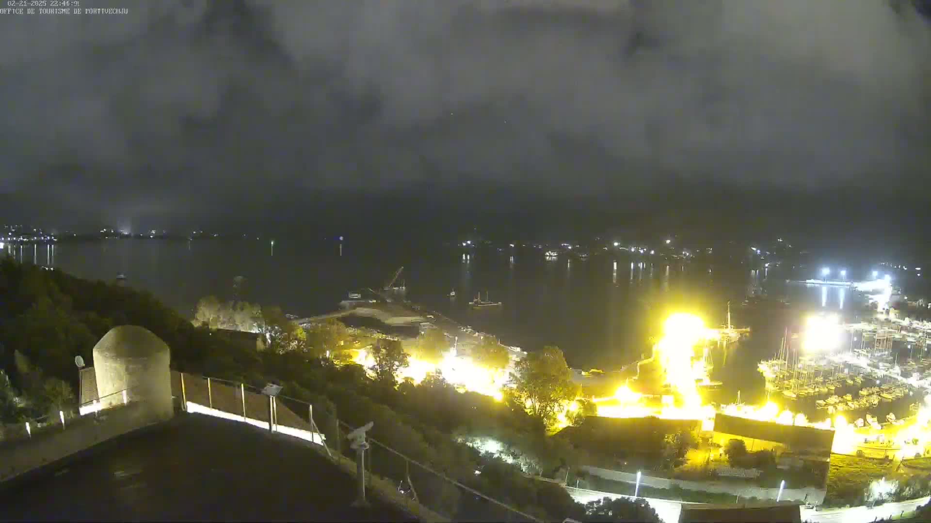 A nighttime, overcast view shows a body of water with a marina and waterfront town, illuminated with artificial lights, seen from an elevated vantage point above a hill.