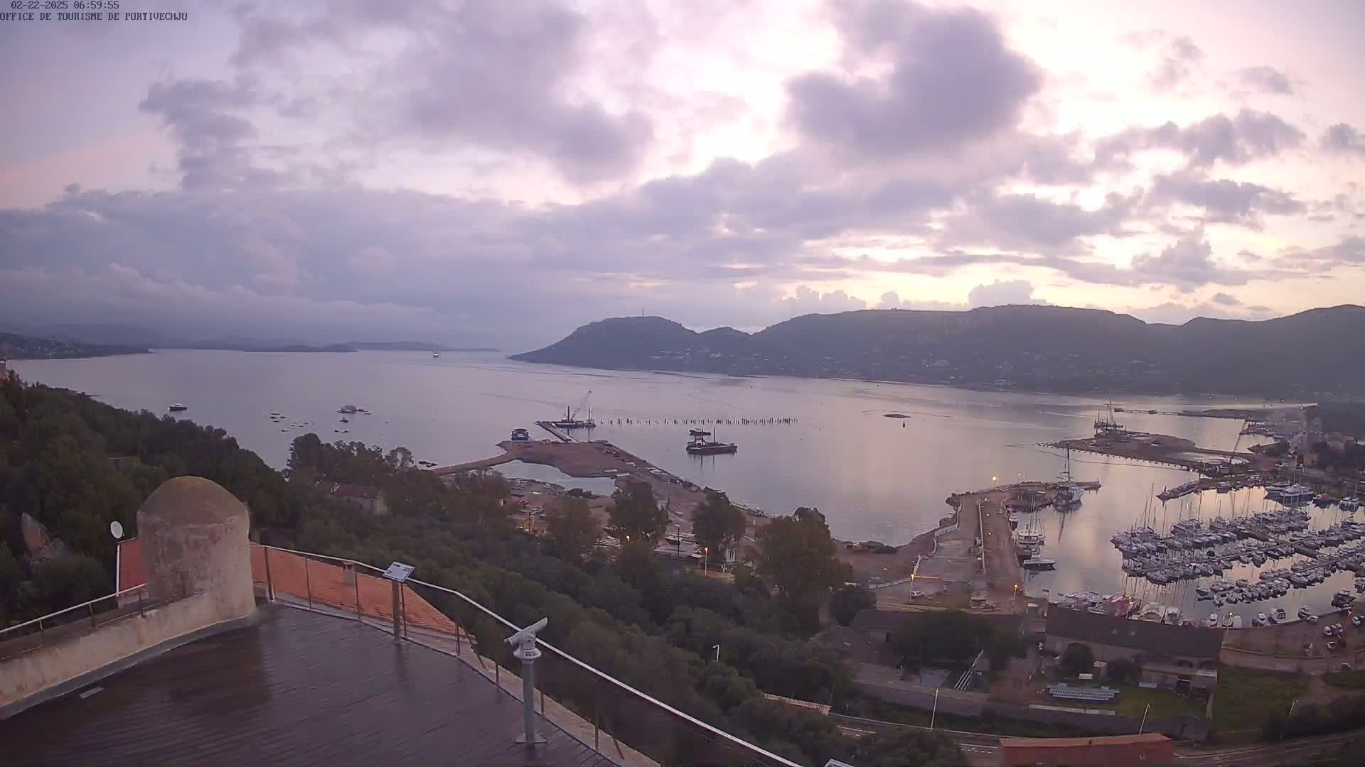 An elevated view shows a calm bay with many boats in a marina, surrounded by hills under a cloudy, dusky sky.