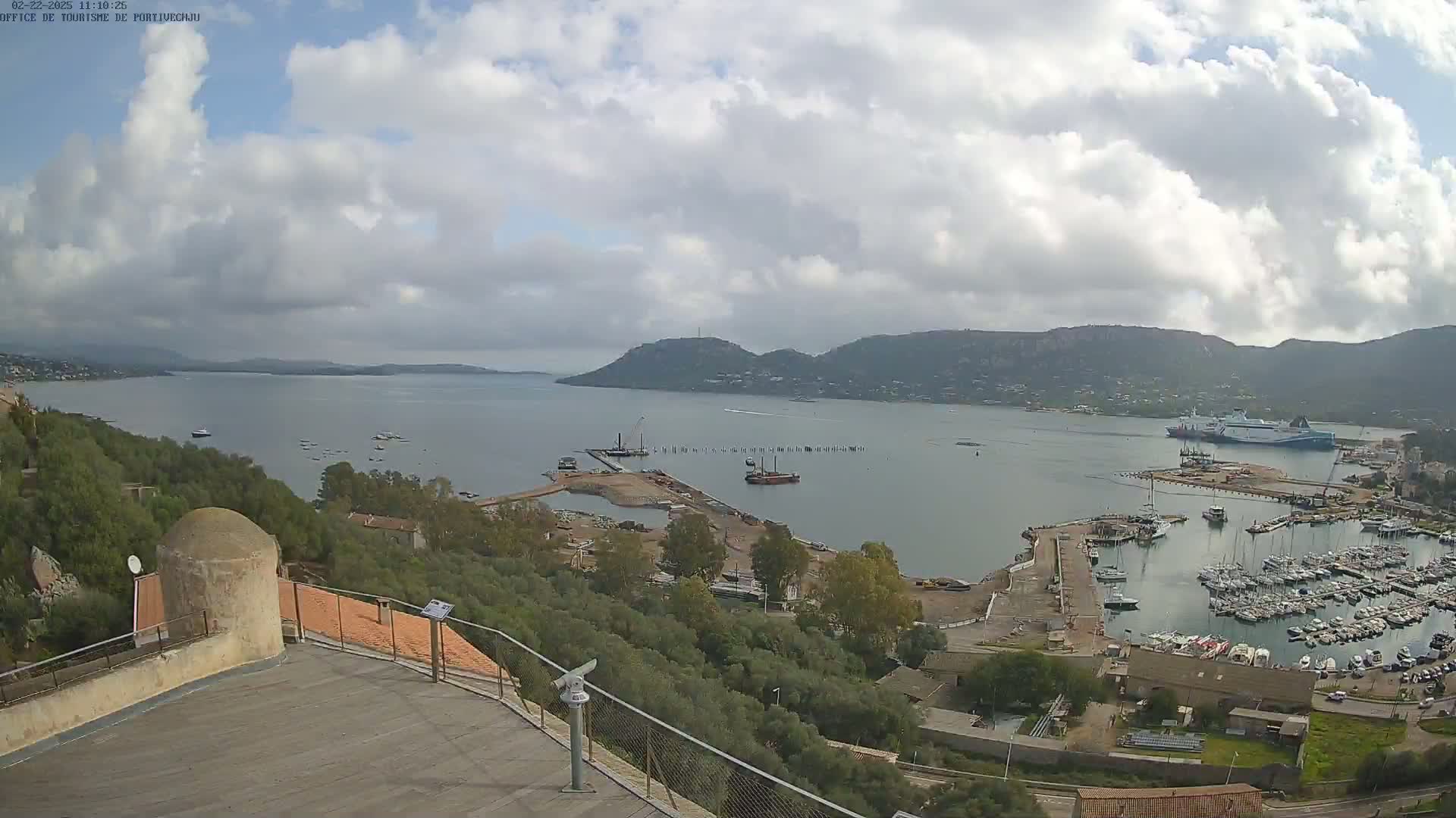 A partly cloudy day overlooks a harbor filled with numerous boats and ships, with hills and a calm bay in the distance.