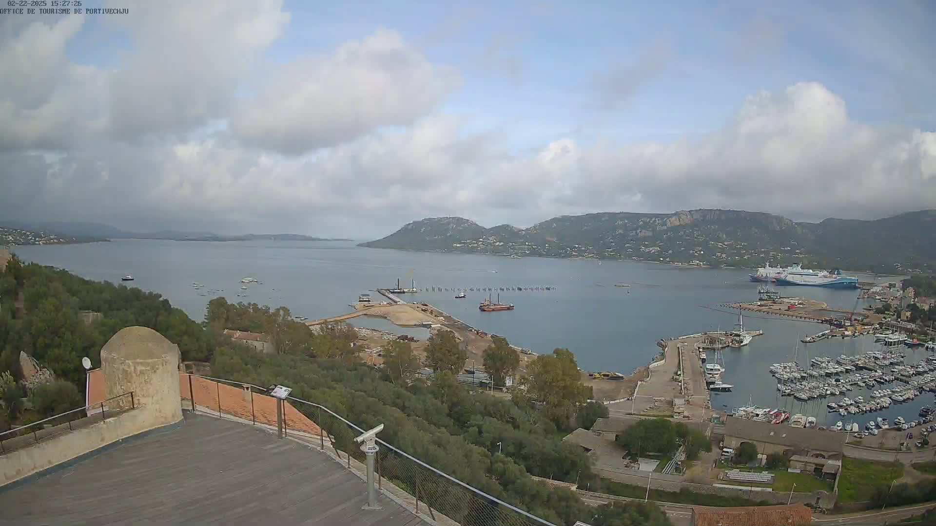 A partly cloudy day overlooks a harbor filled with boats, a large ship, and ongoing construction, with hills and water in the background.