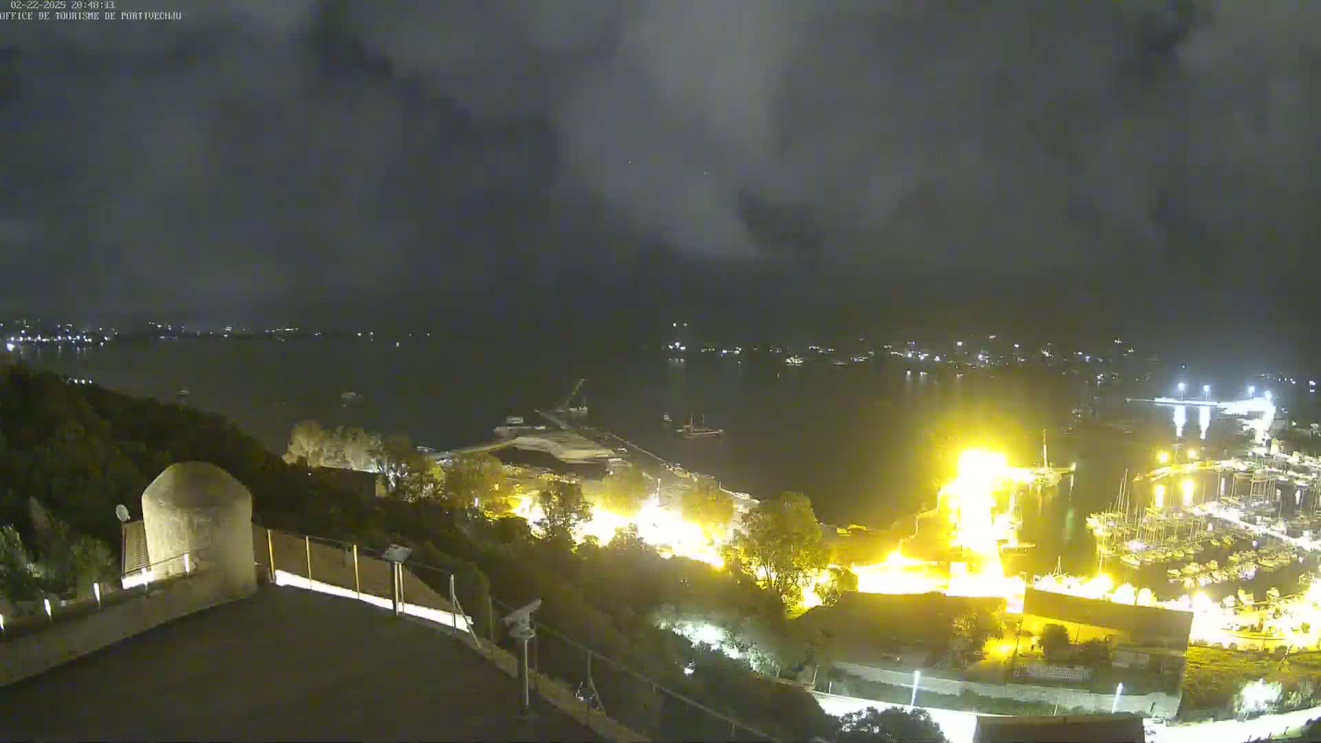 A nighttime view from an elevated position shows a harbor with brightly lit boats and buildings, under a cloudy sky.