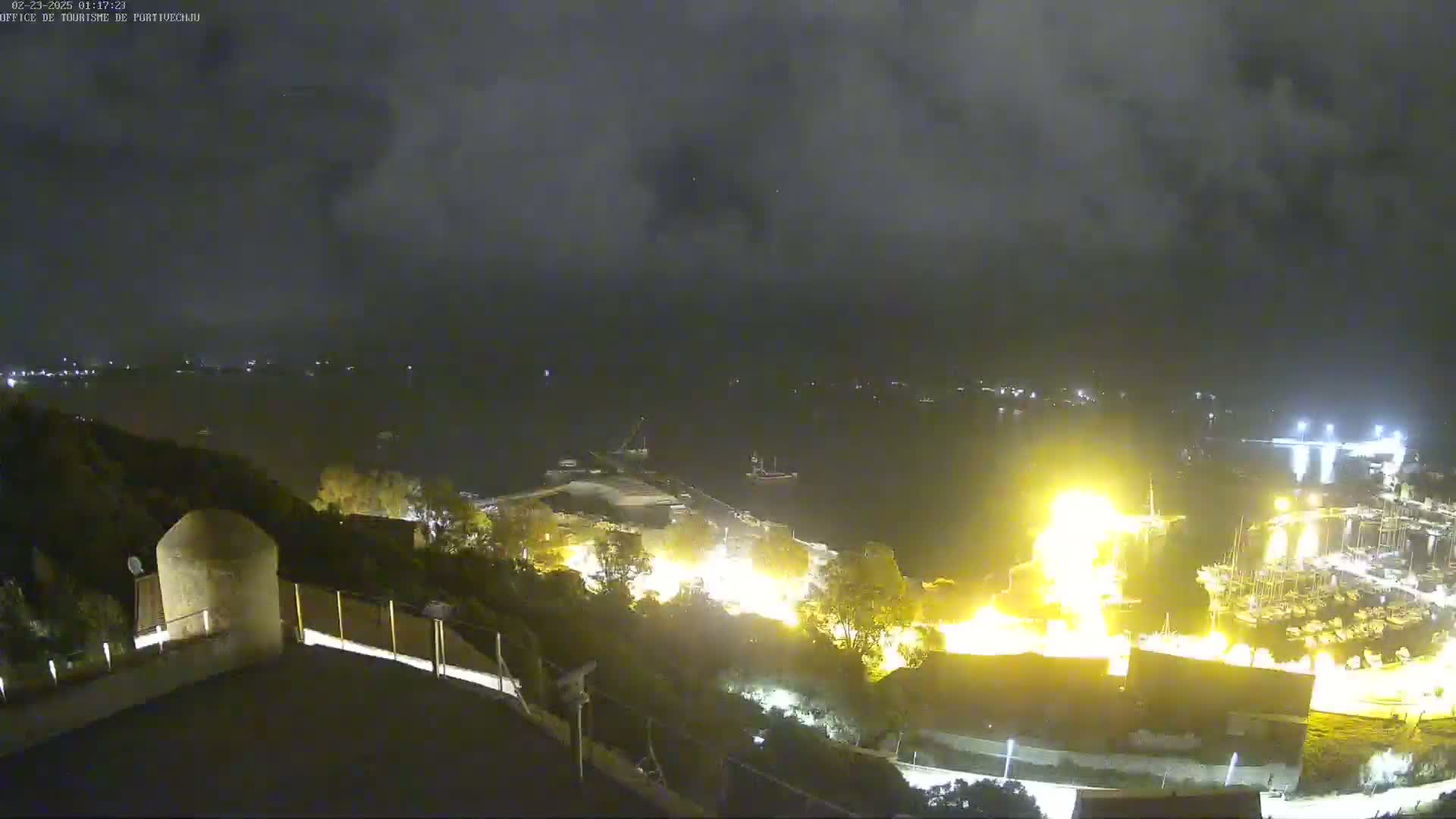 A nighttime, high-angle view shows a body of water with a brightly lit area near a shoreline, possibly a fire or construction site, under a cloudy sky.