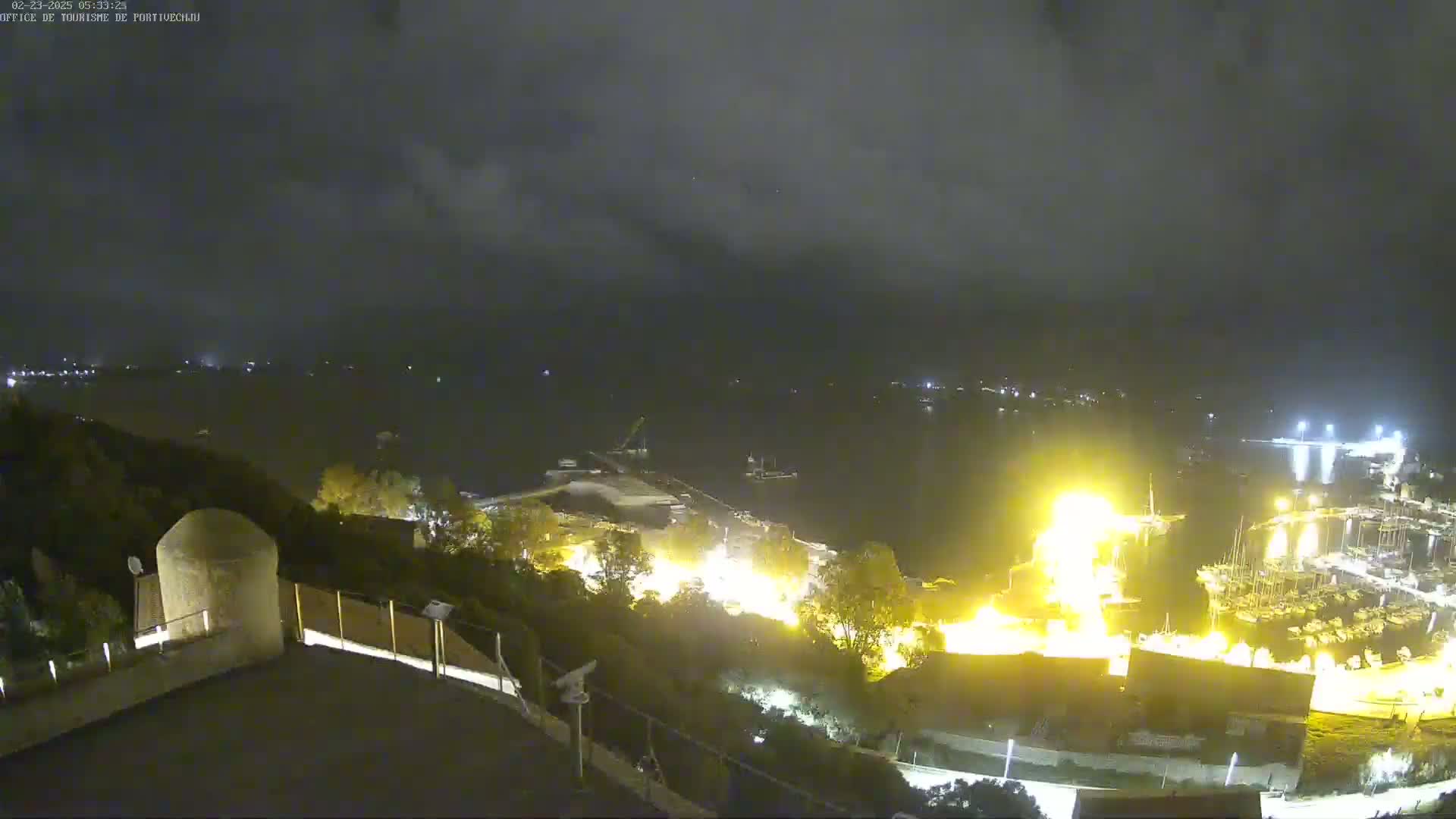 A nighttime, cloudy view from an elevated position overlooking a harbor with brightly lit boats and buildings, a large bright area suggesting a fire or intense light source, and dark water.