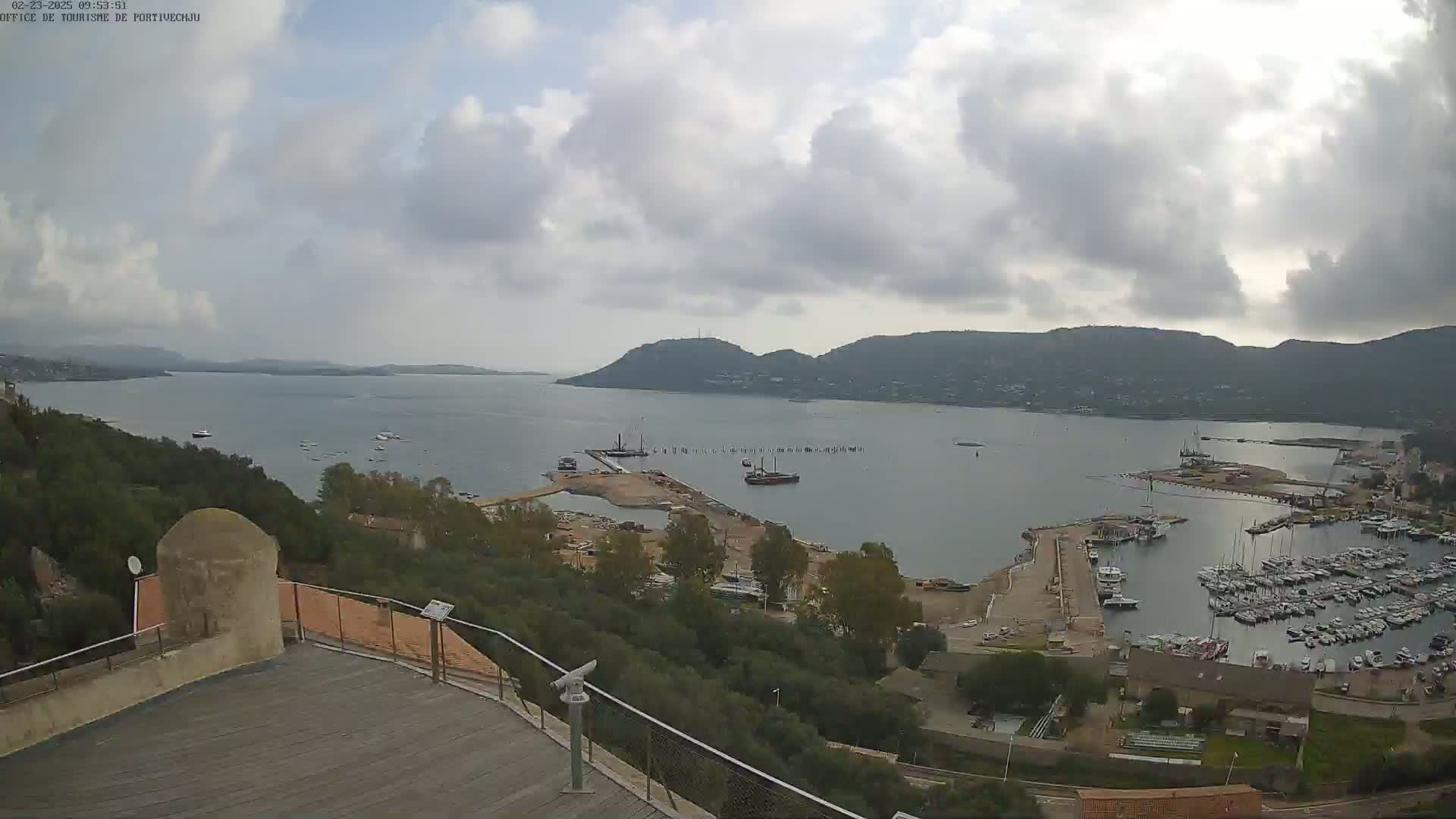 An elevated view of a harbor filled with boats on a cloudy day, with hills and a calm body of water in the background.
