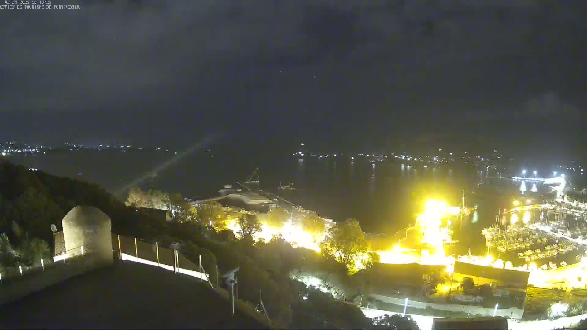 A nighttime, high-angle view of a harbor filled with boats, illuminated by bright lights, with a dark, cloudy sky above and a hilly landscape in the foreground.