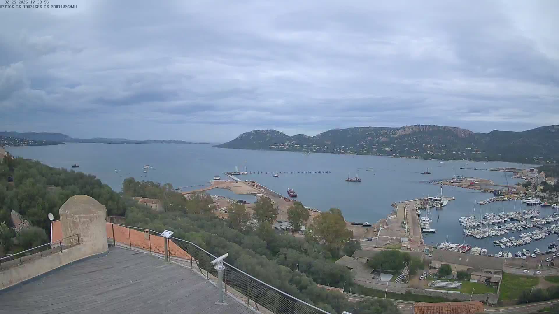 A high-angle view of a harbor filled with boats, under a mostly cloudy sky, with hills in the background.