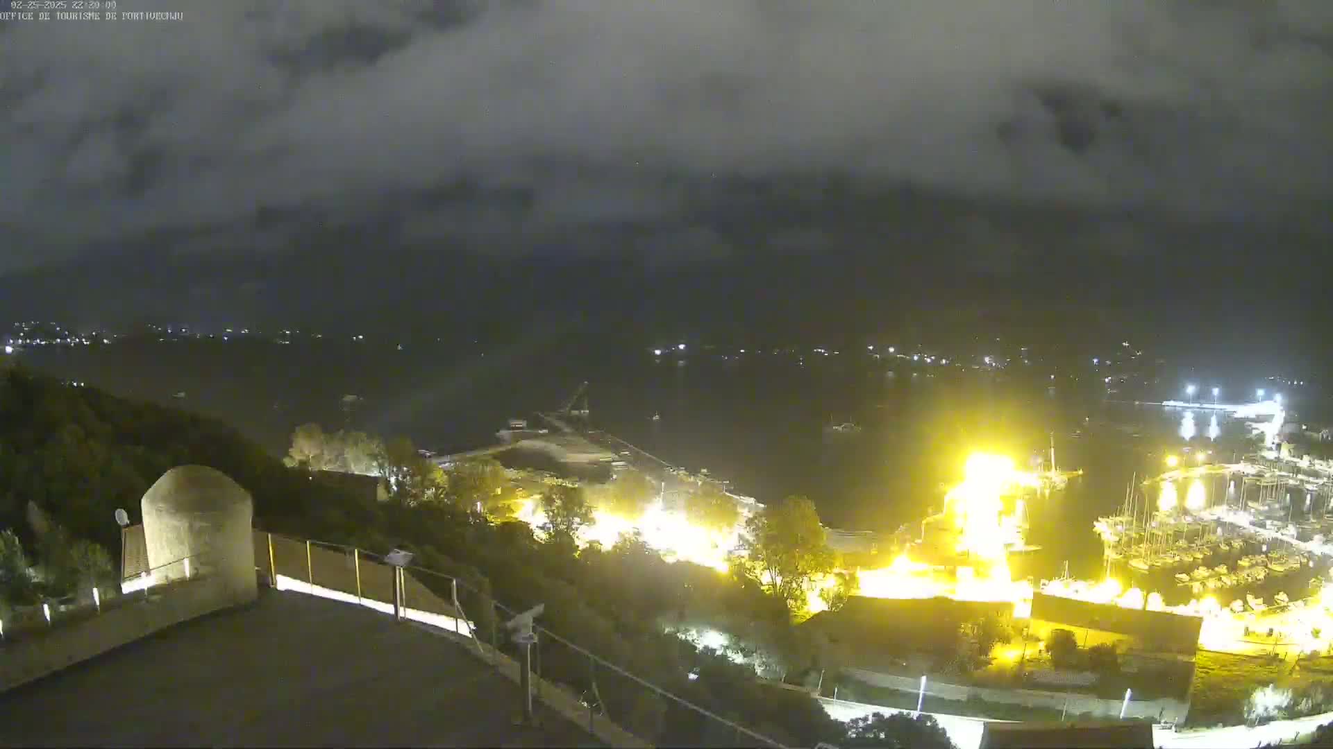 A nighttime view from an elevated vantage point shows a harbor with bright lights and a large fire, under a dark, cloudy sky.