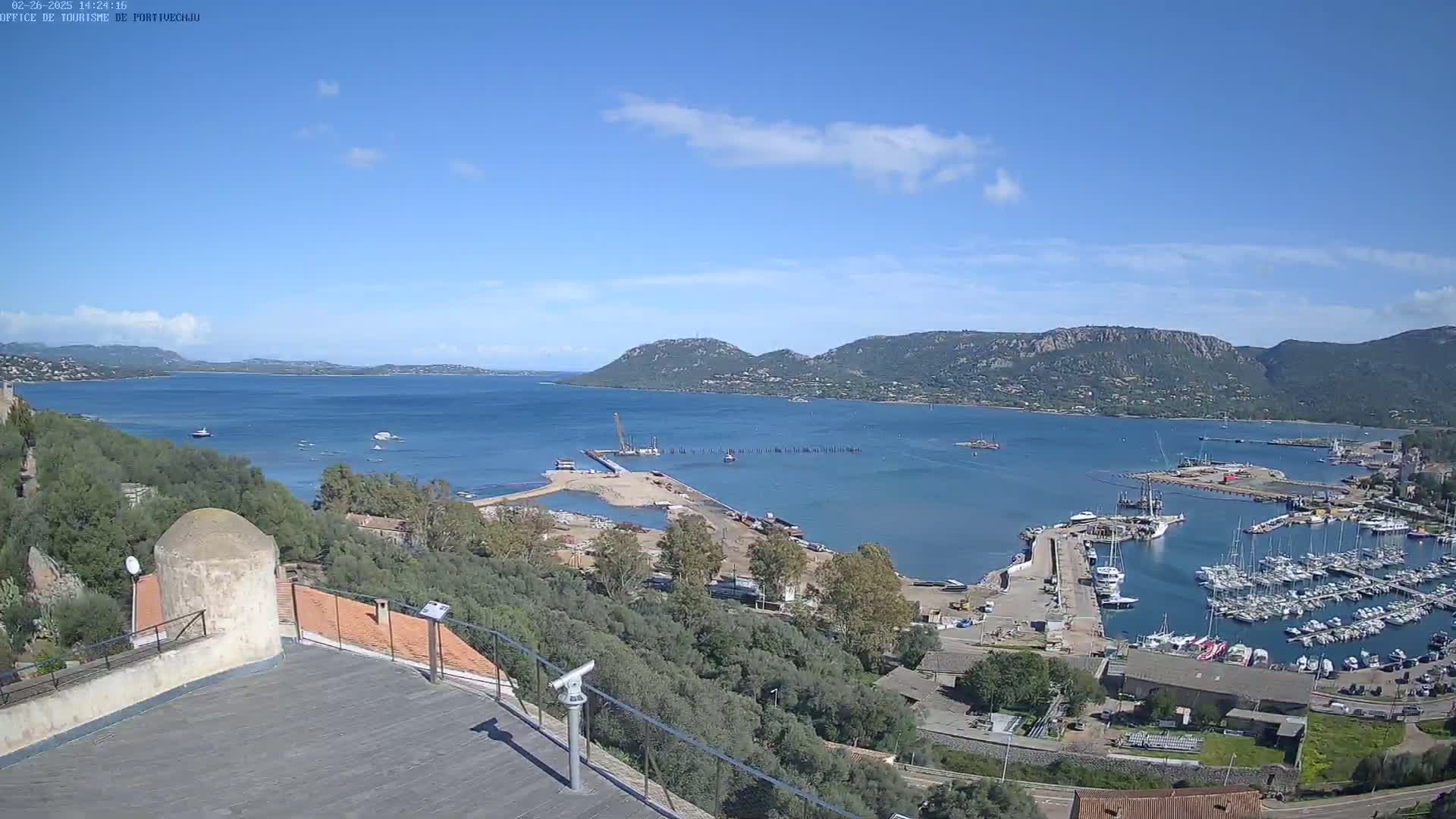 A high-angle view of a harbor filled with boats, under a mostly clear sky, with construction visible near the harbor entrance and hills in the distance.