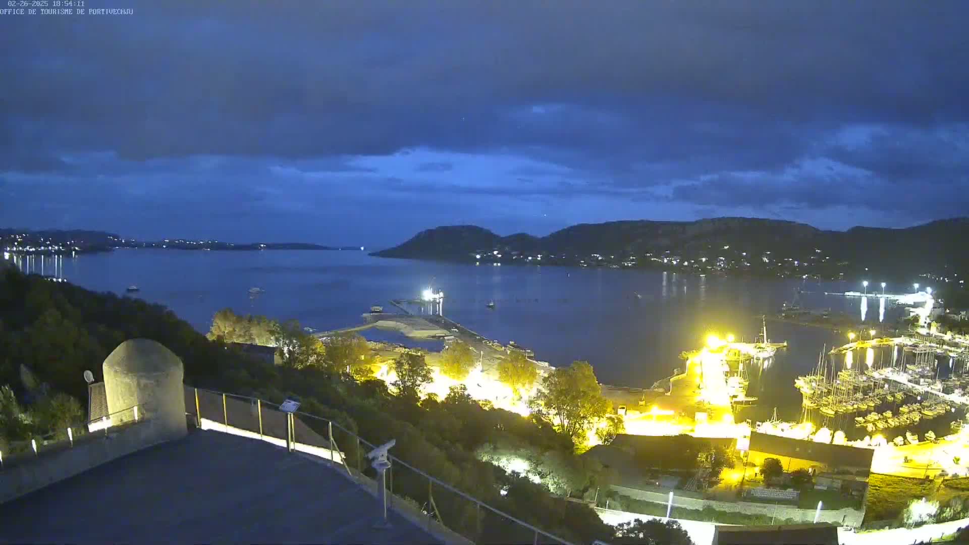 A nighttime view of a harbor filled with boats, lit by artificial lights under a partly cloudy, dark blue sky.