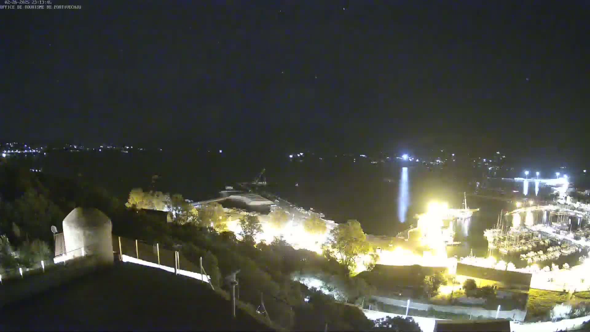 A nighttime, high-angle view of a harbor with boats docked, illuminated buildings on a hillside, and a clear, starry sky.