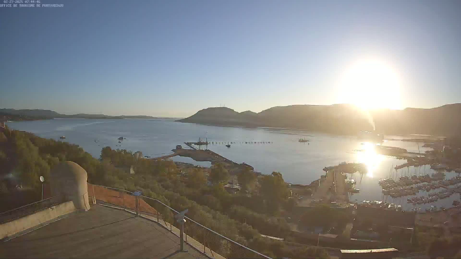 A high-angle, sunny view overlooking a calm bay with a marina filled with boats, a small town nestled along the shoreline, and hills in the distance.
