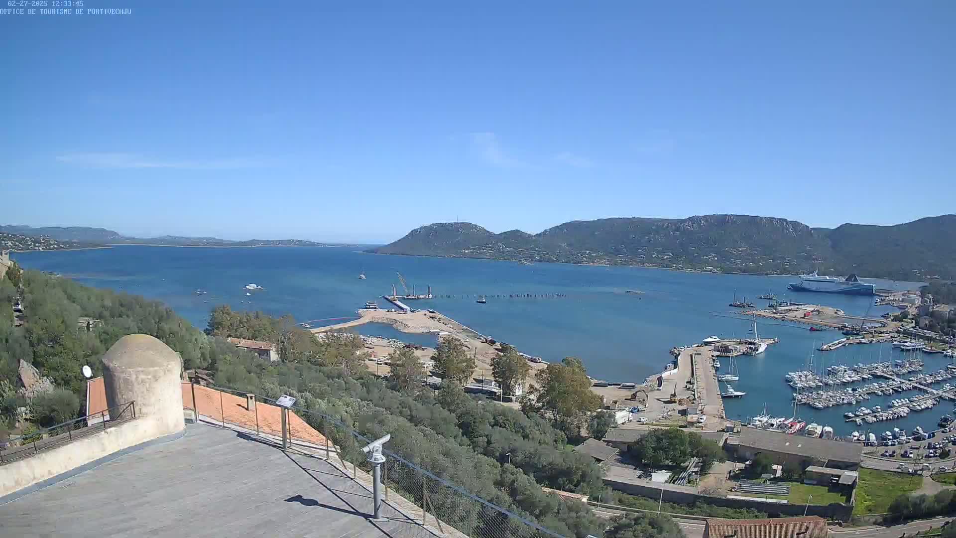 A panoramic view of a harbor filled with boats, under a clear blue sky, shows ongoing construction activity near a calm body of water surrounded by hills.