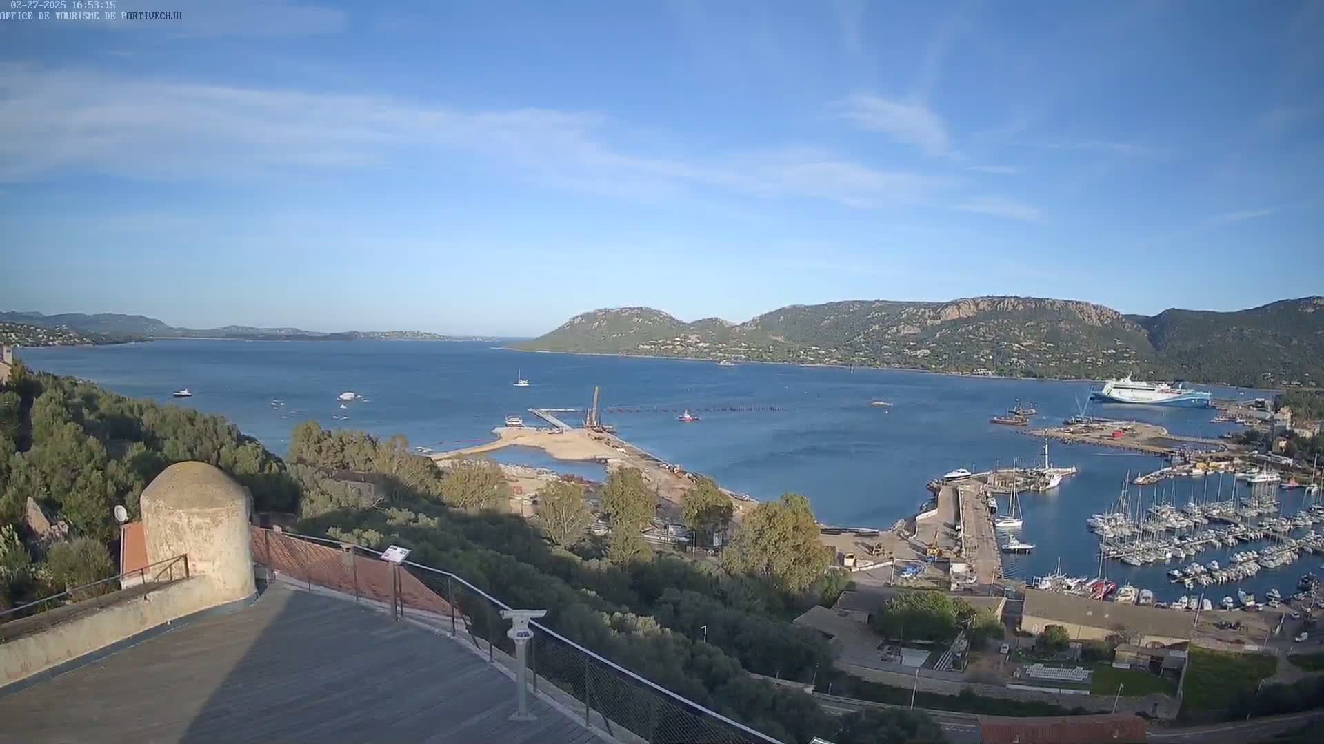 A high-angle, sunny view shows a calm bay with numerous boats and a large ferry, construction activity near a sandy shoreline, and hills in the background.