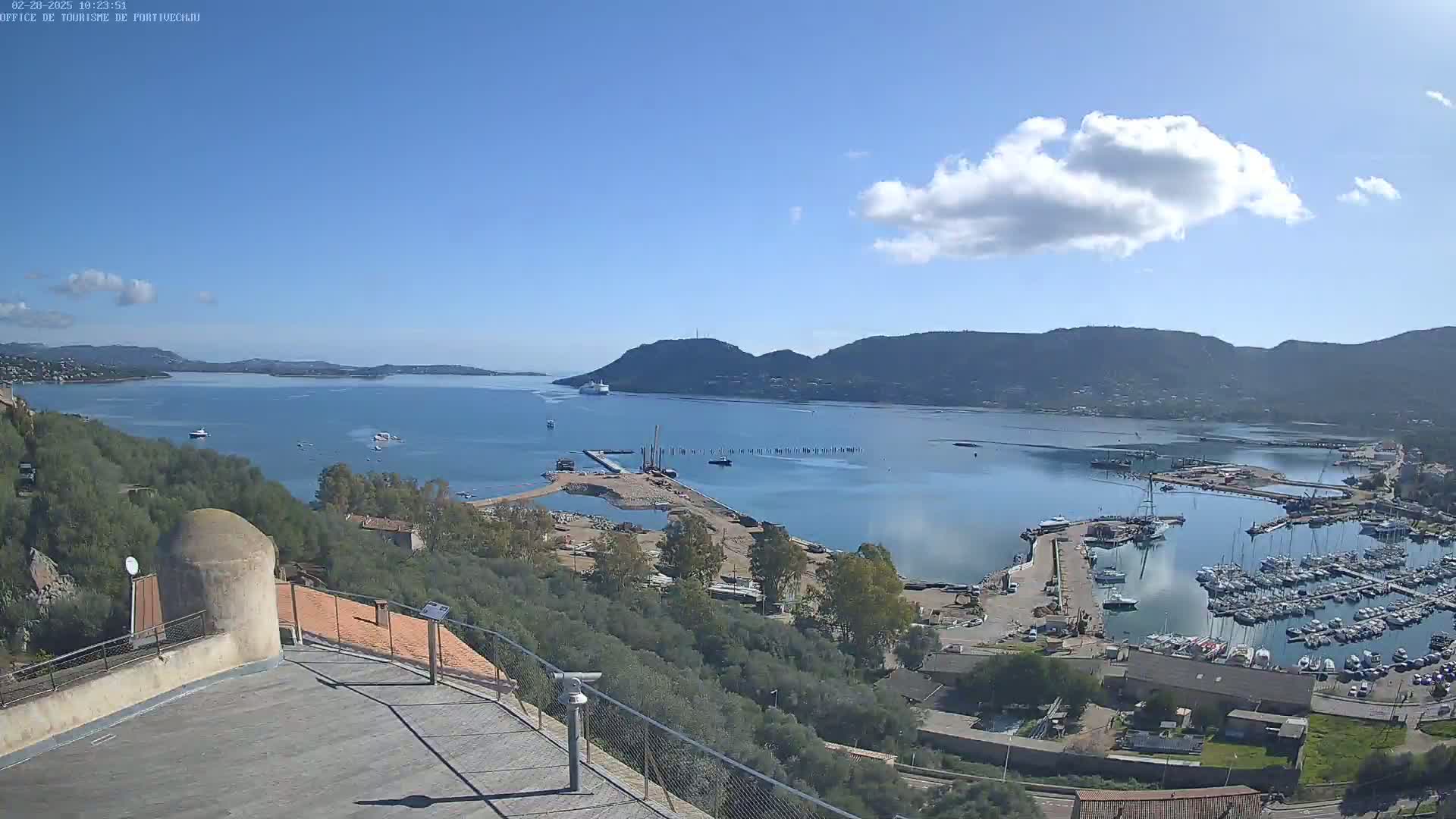 A high-angle, sunny view of a harbor filled with boats, construction equipment, and buildings nestled in a bay surrounded by hills under a mostly clear blue sky with a single, puffy cloud.