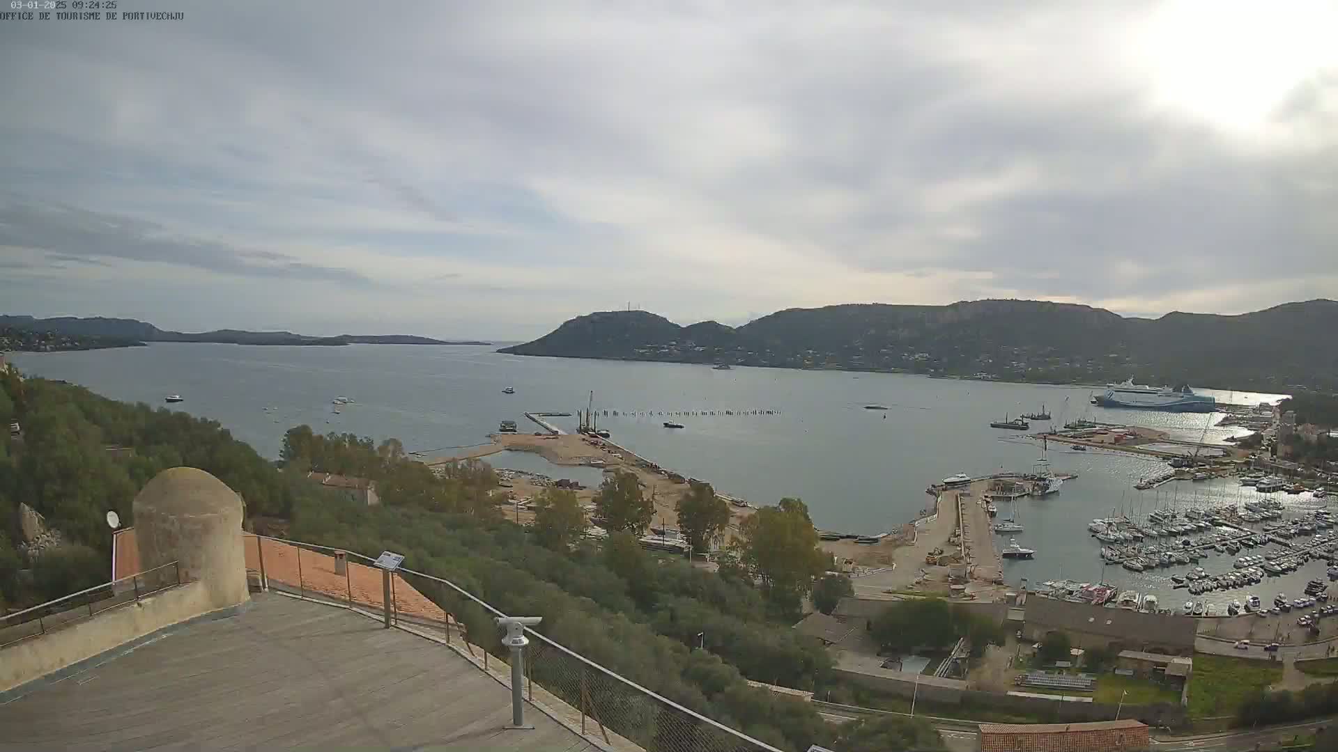 A high-angle view shows a harbor filled with numerous boats and ships on a partly cloudy day, with hills and a body of water visible in the background.