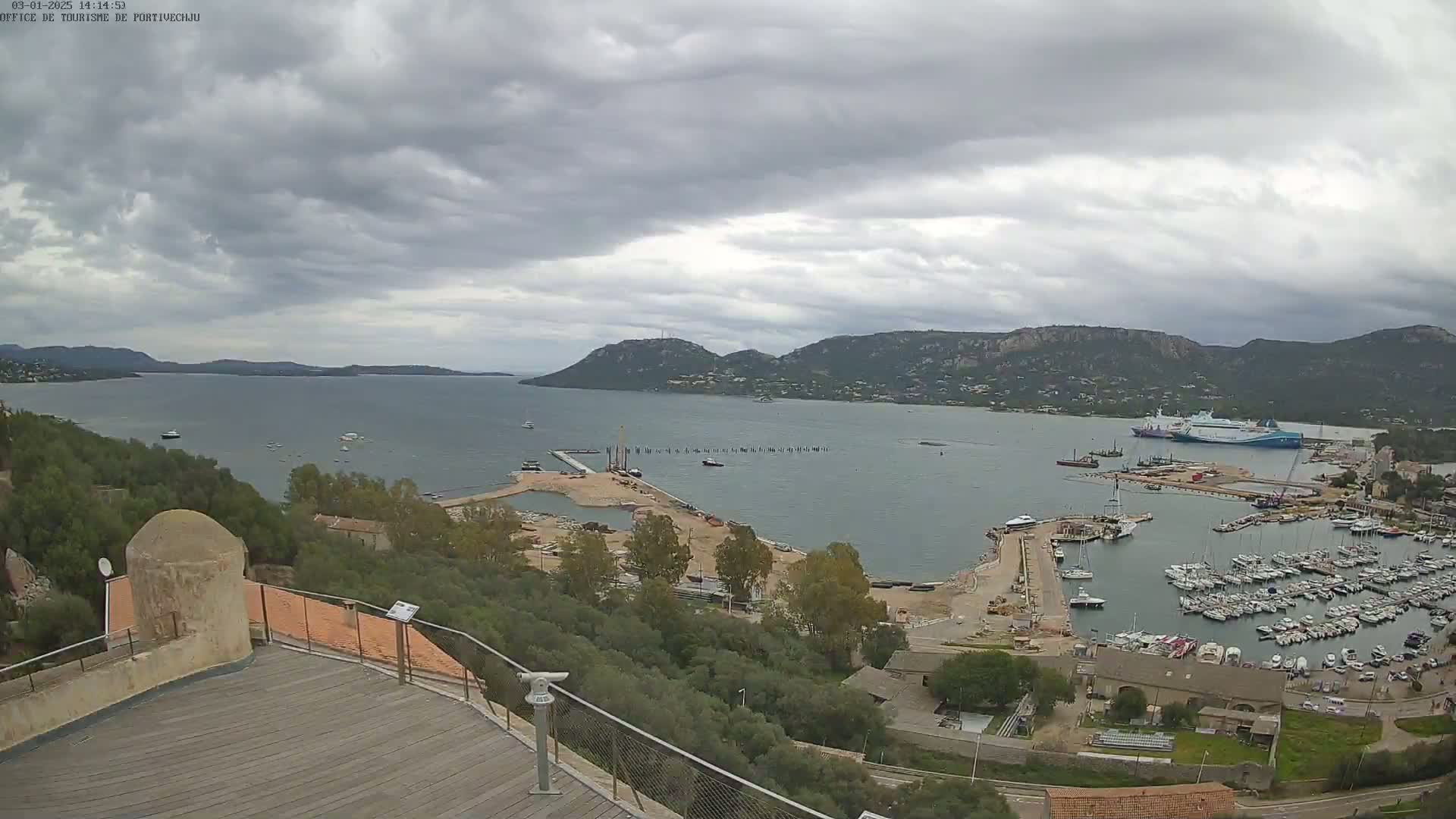 An elevated view shows a harbor filled with numerous boats and ships under a cloudy sky, with hills in the background.