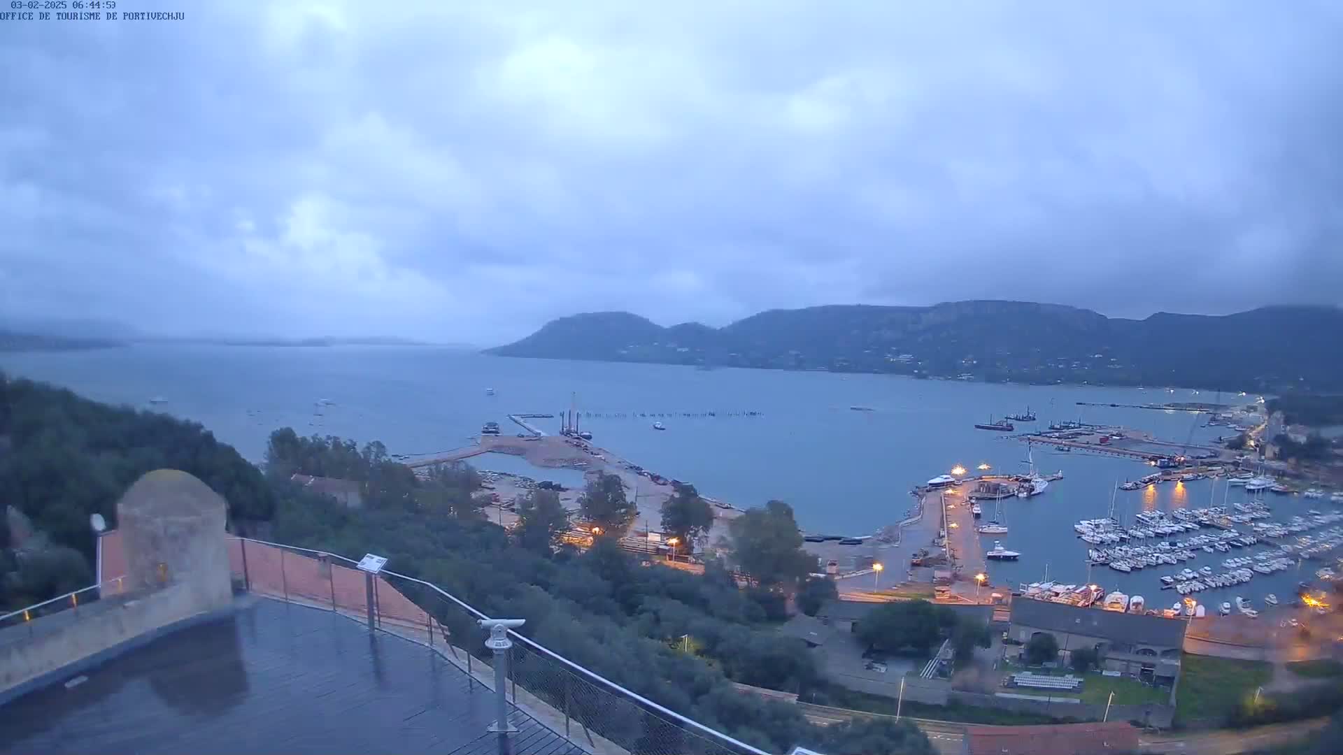 A harbor filled with boats is nestled in a bay surrounded by hills under an overcast sky.