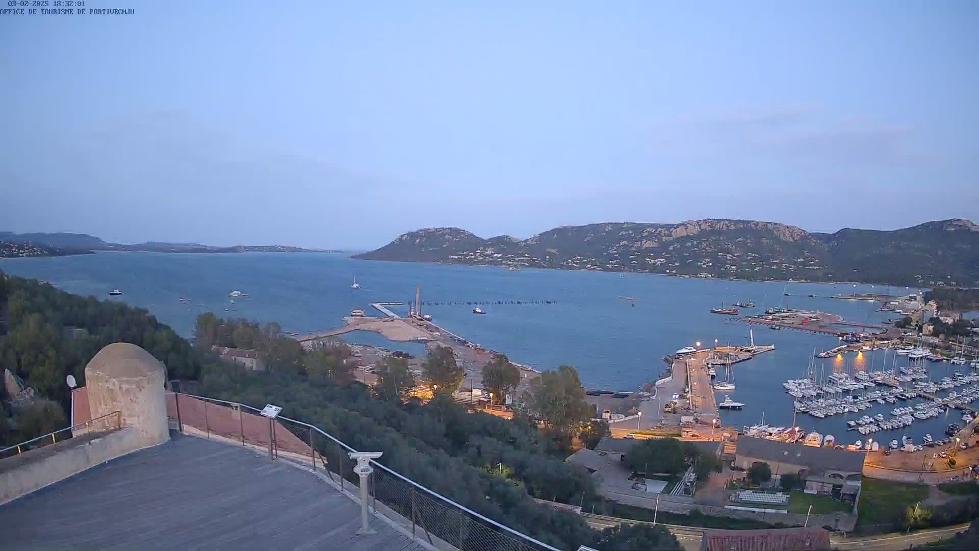 A high-angle, twilight view of a harbor filled with numerous boats, situated next to a town nestled against a mountainous coastline under a clear sky.