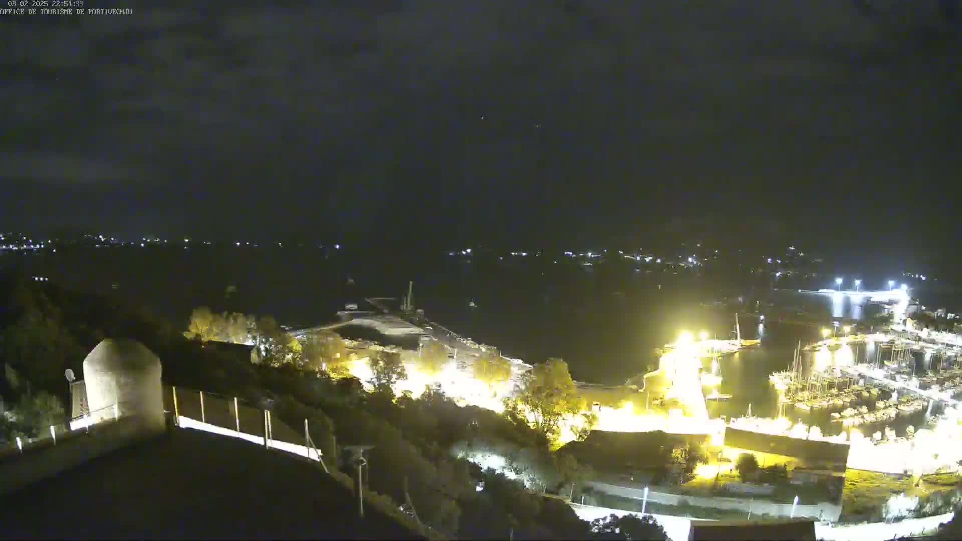A nighttime, high-angle view of a harbor filled with boats, illuminated by lights, with a hilltop overlooking the scene.