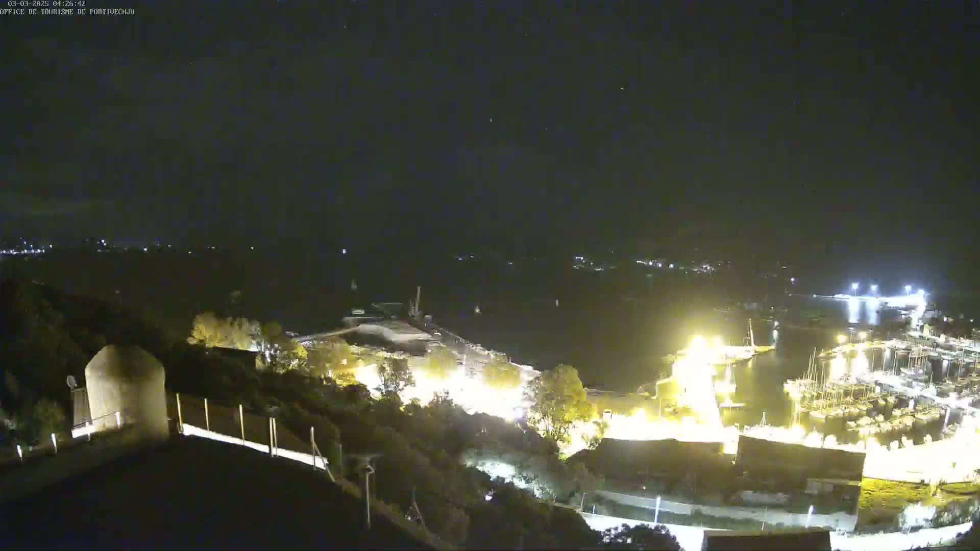 A nighttime, high-angle view shows a harbor filled with boats, illuminated by artificial lights, with a dark, hilly area in the foreground.