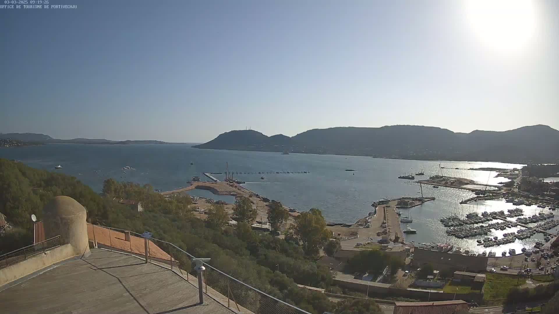 A sunny day overlooking a harbor filled with boats, a construction area, and a calm bay surrounded by hills.