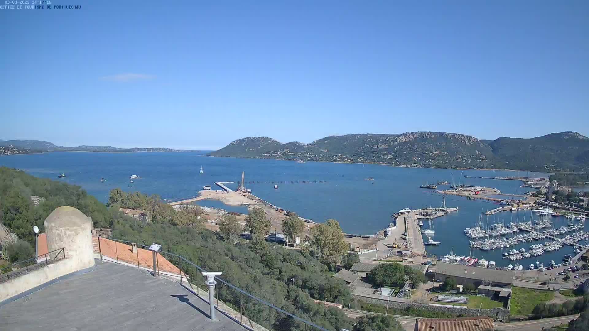 A high-angle, long shot on a clear, sunny day shows a calm bay with numerous boats in a marina, construction activity near the water, and hills in the background.