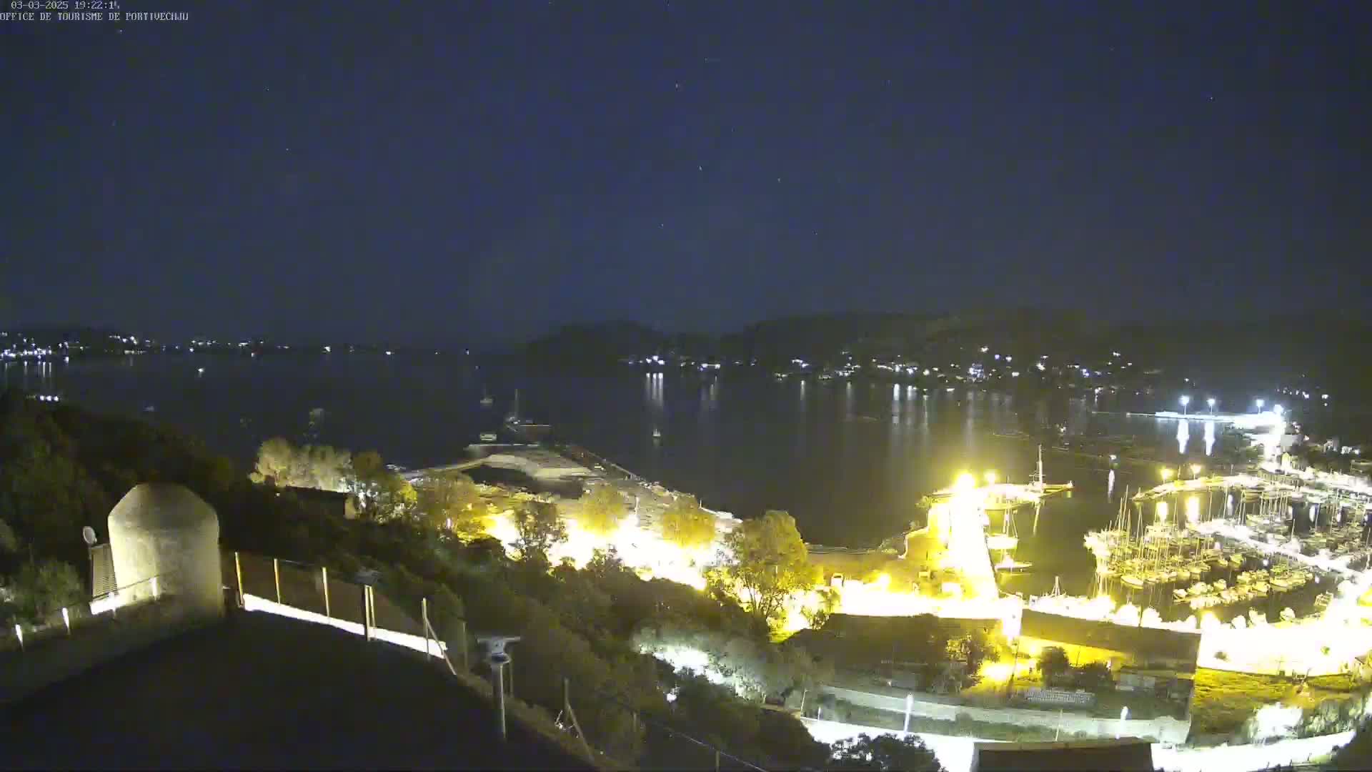 A nighttime view of a harbor filled with boats, illuminated by lights, under a clear, starry sky.