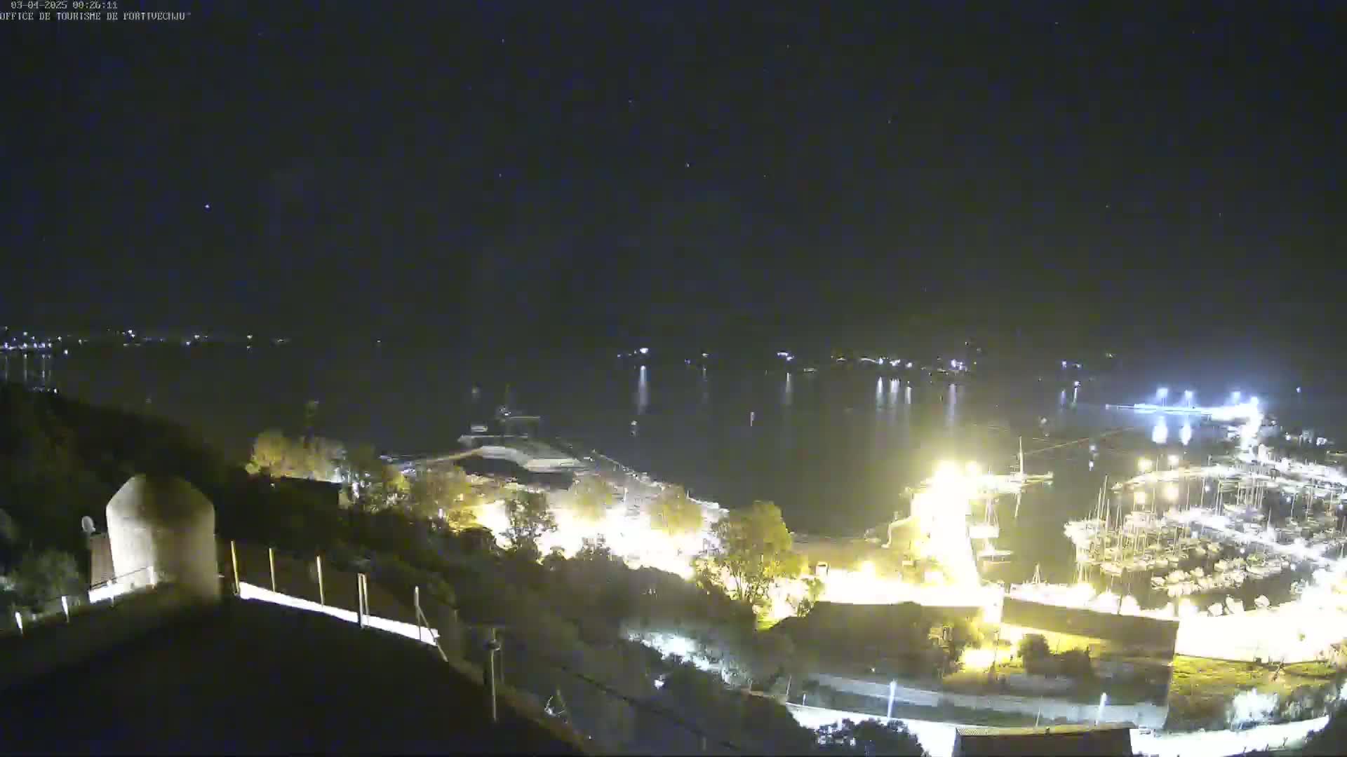 A nighttime, high-angle view shows a harbor filled with boats, illuminated by lights against a dark sky with some stars visible.