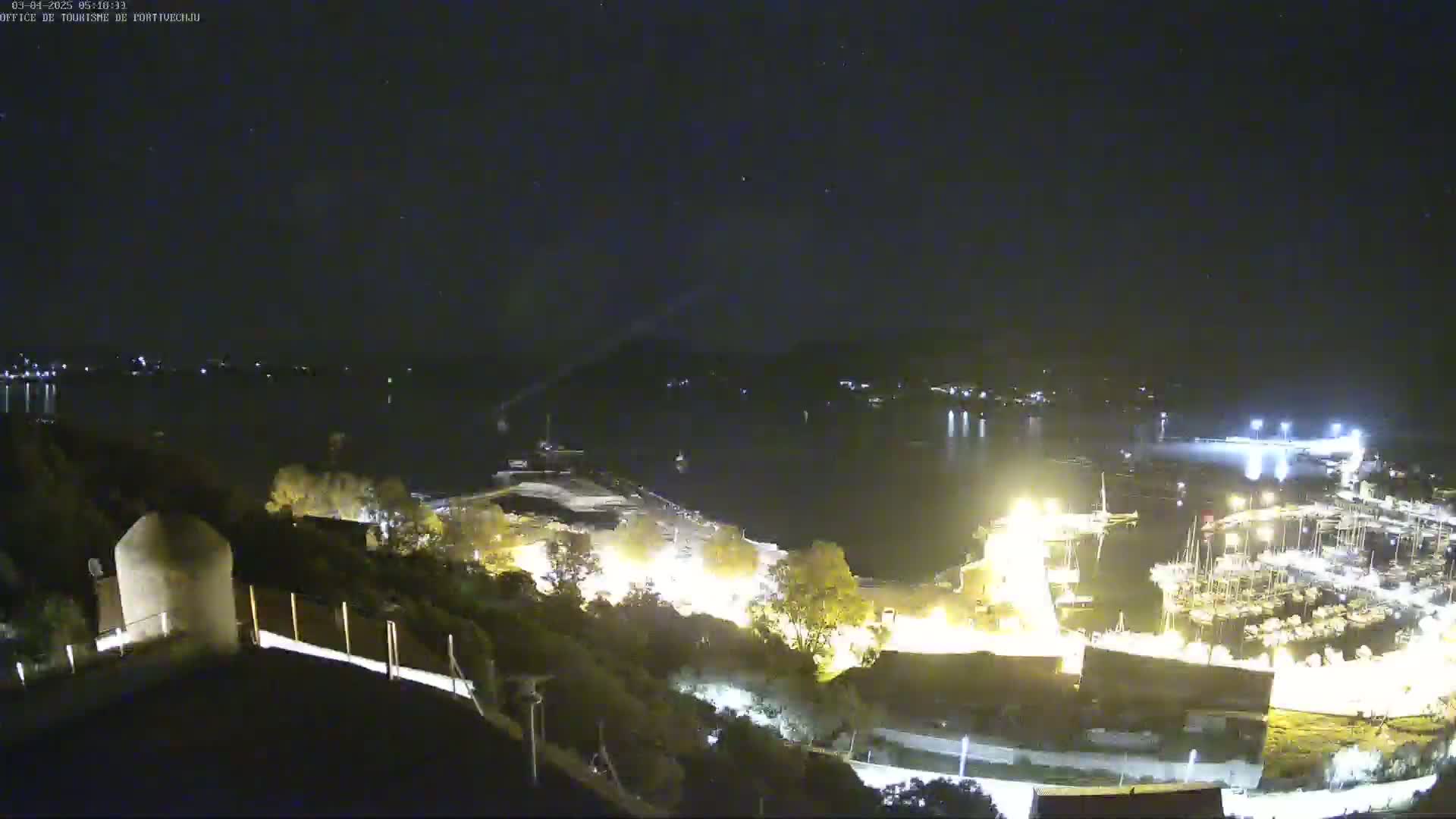 A nighttime view from an elevated position shows a calm body of water with a marina filled with boats, and a town illuminated with lights under a clear, starry sky.